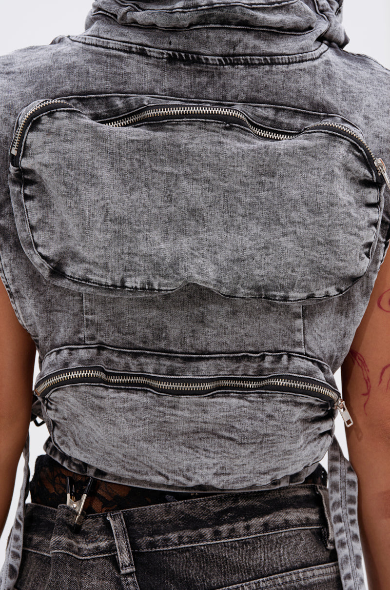 A close-up of a person wearing the MODEL BEHAVIOR DENIM TOP, a sleeveless cropped acid-wash black vest with two large zippered back pockets. The person, showing tattooed arms, carries a matching denim bag.