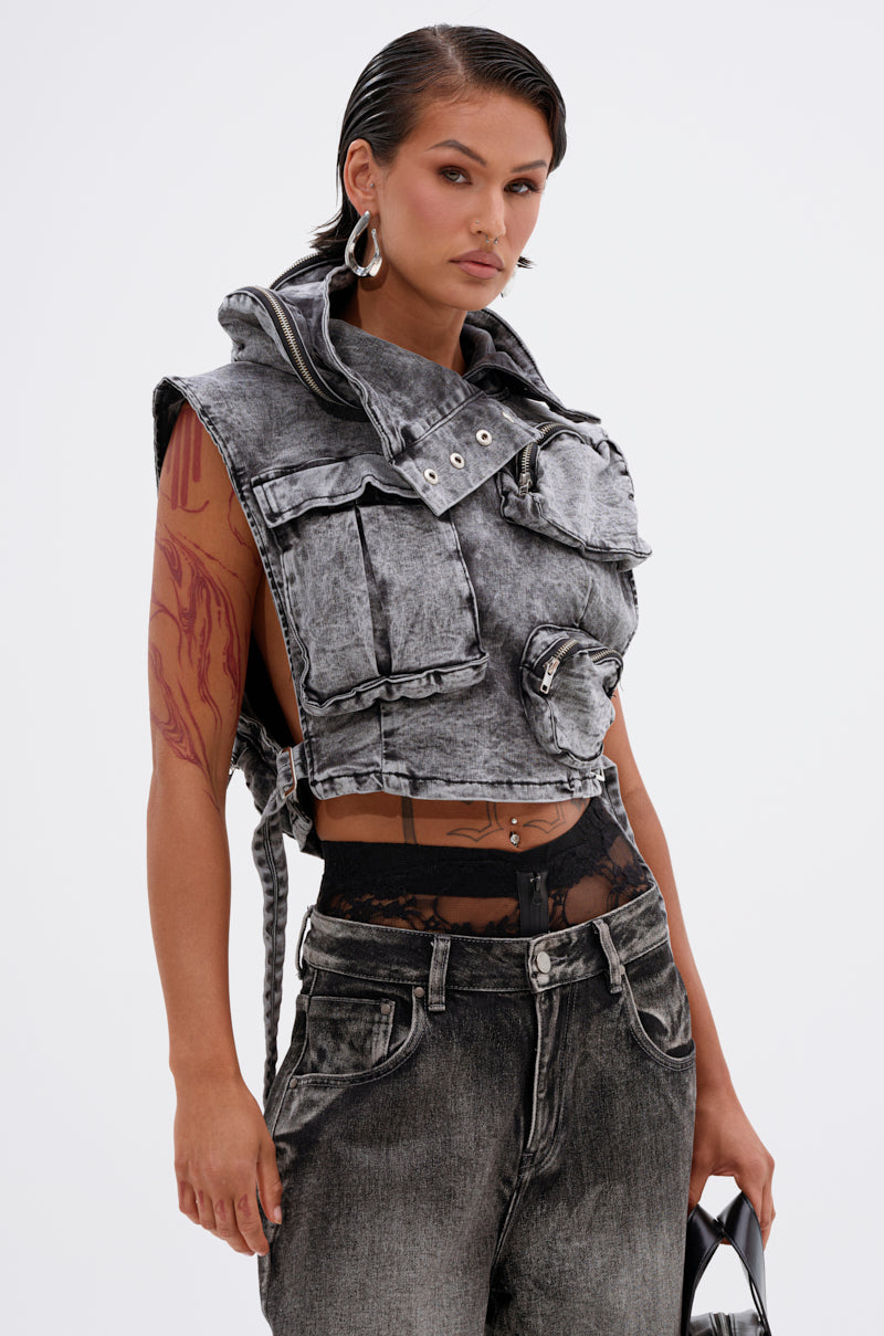 A woman with wet-look hair wears the cropped MODEL BEHAVIOR DENIM TOP in distressed gray with oversized pockets, paired with matching gray jeans. She accessorizes with large hoop earrings, a black handbag, and lace underwear peeking above her jeans.