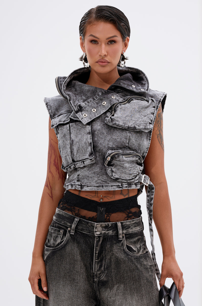 A woman with slicked-back hair wears the MODEL BEHAVIOR DENIM TOP, a sleeveless cropped acid-wash black denim vest with multiple pockets, layered over a black lace top and matching acid-washed jeans. She holds a bag against a plain white background.