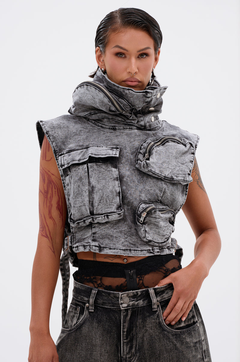 A person with slicked-back hair wears the MODEL BEHAVIOR DENIM TOP, a cropped, acid-wash black vest with large pockets and a high collar, paired with matching jeans. A red tattoo is visible on one arm and black lace peeks out at the waist.