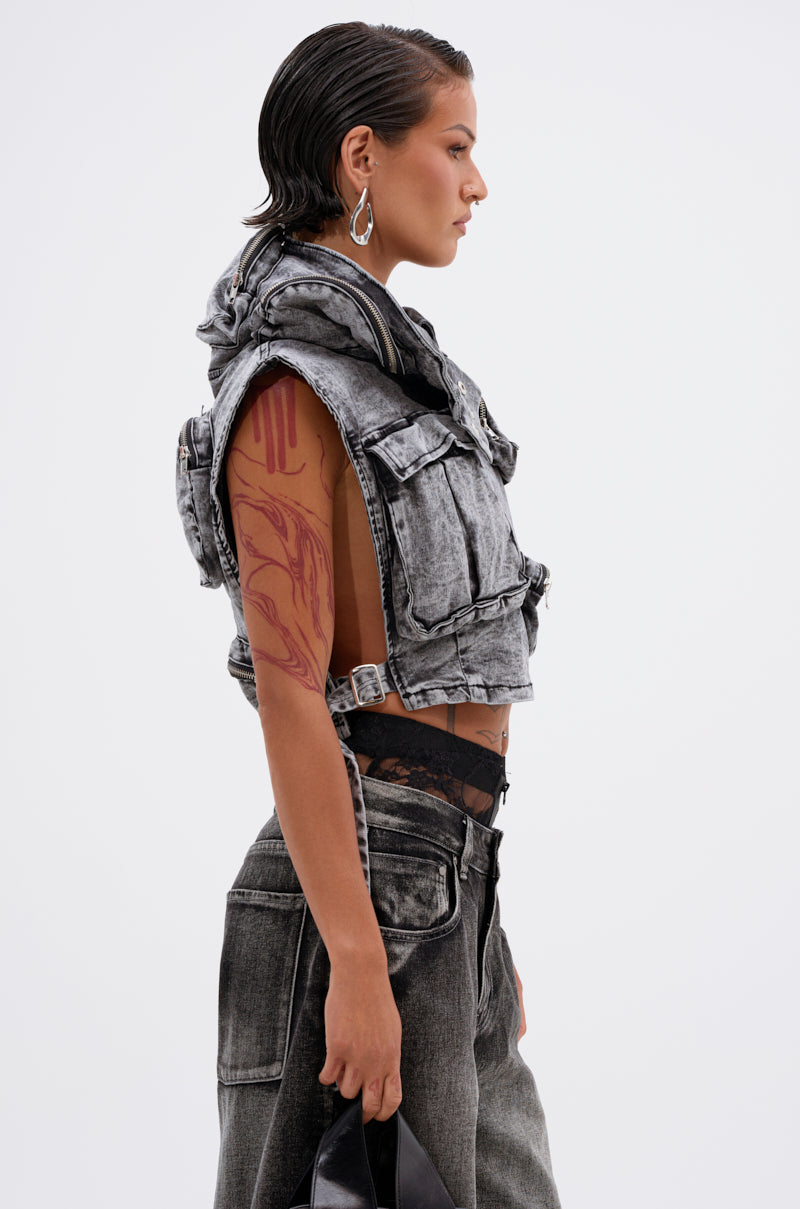 A person with short, slicked-back hair wears the MODEL BEHAVIOR DENIM TOP, a sleeveless cropped acid-wash black vest, paired with matching jeans. They show arm tattoos, large geometric earrings, and hold a black bag at their side.