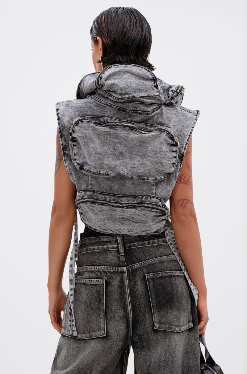 A person with wet, slicked-back hair wears the MODEL BEHAVIOR DENIM TOP, a cropped acid-wash black vest with a large collar, paired with matching wide-leg jeans. The photo is shot from behind, revealing tattoos on their left arm.