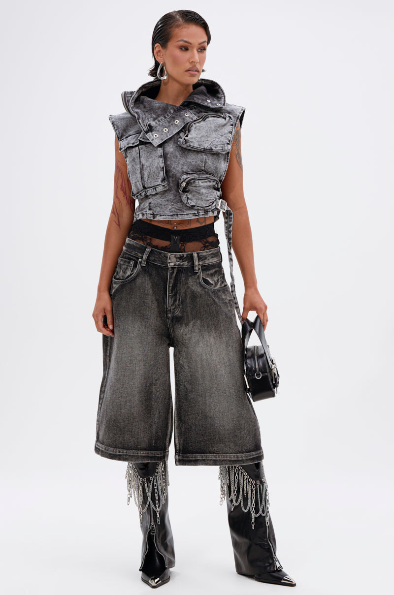 A woman wears the MODEL BEHAVIOR DENIM TOP, paired with wide-leg acid-wash black denim shorts, shiny black boots with chain fringe, and holds a small black bag, posing against a plain white background.