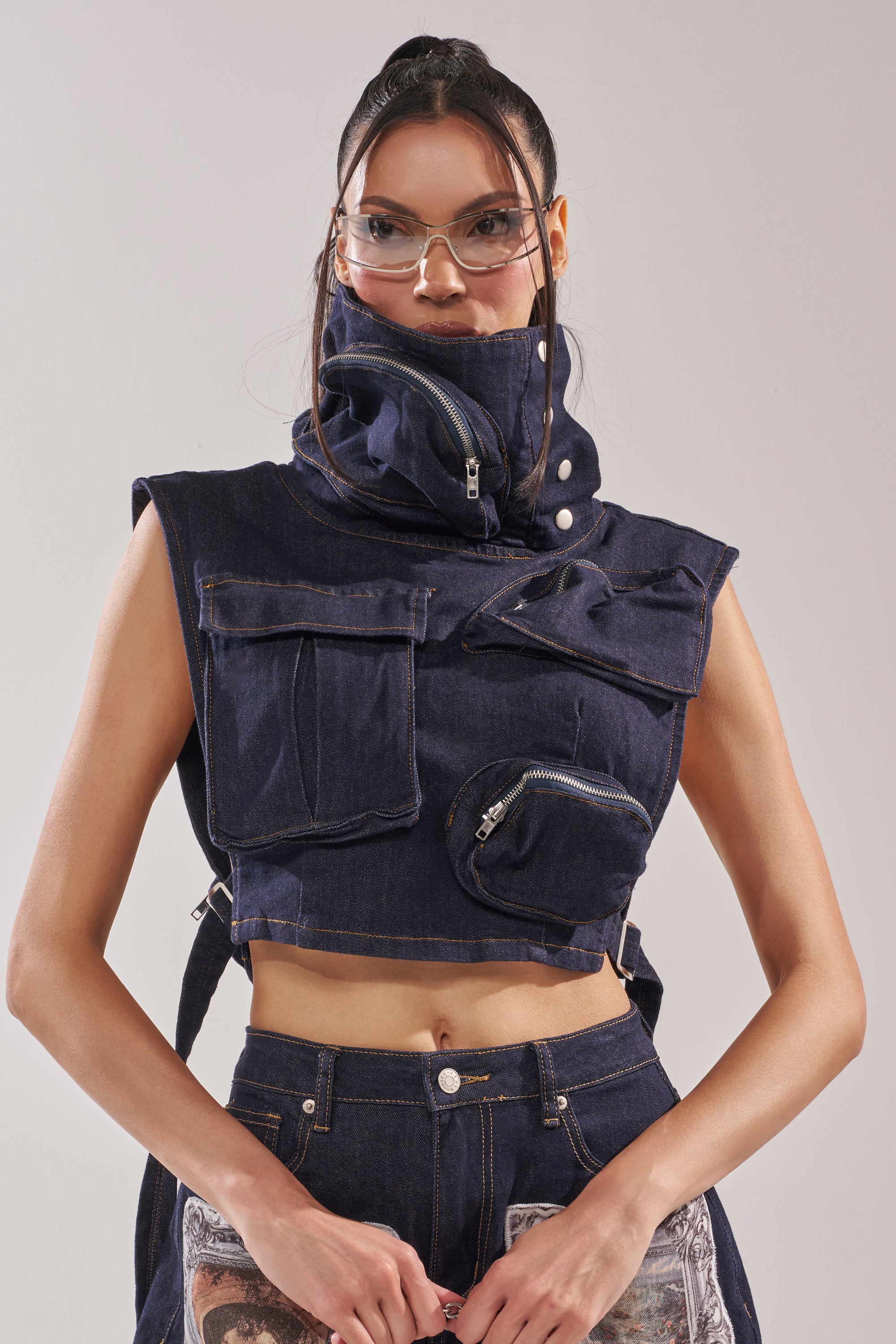 A woman wears the MODEL BEHAVIOR TOP IN DARK BLUE DENIM, paired with matching high-waisted jeans and clear protective glasses, as she poses against a plain backdrop. 0