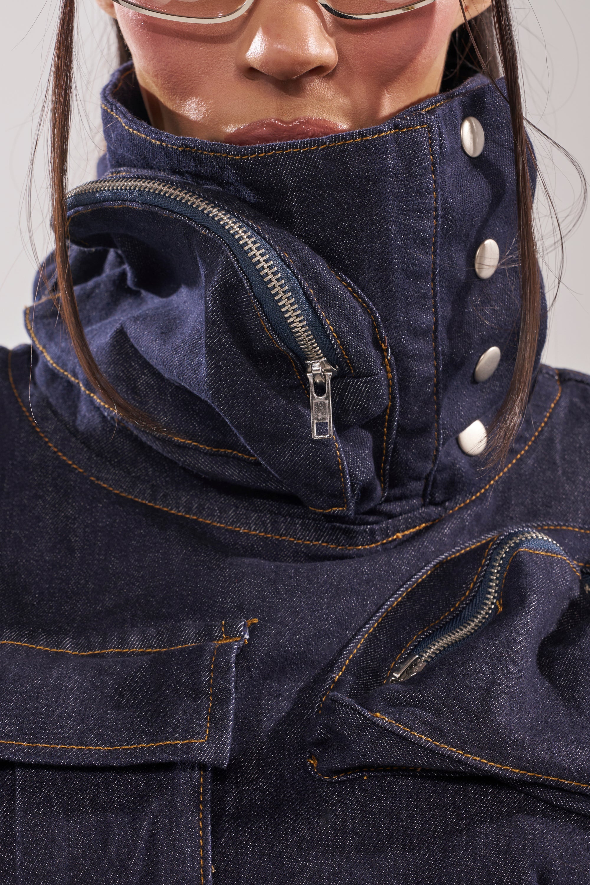Wearing the MODEL BEHAVIOR TOP IN DARK BLUE DENIM with a high collar, large silver buttons, and visible zipper partially zipped to cover the lower face; clear-frame glasses complete the look. 9