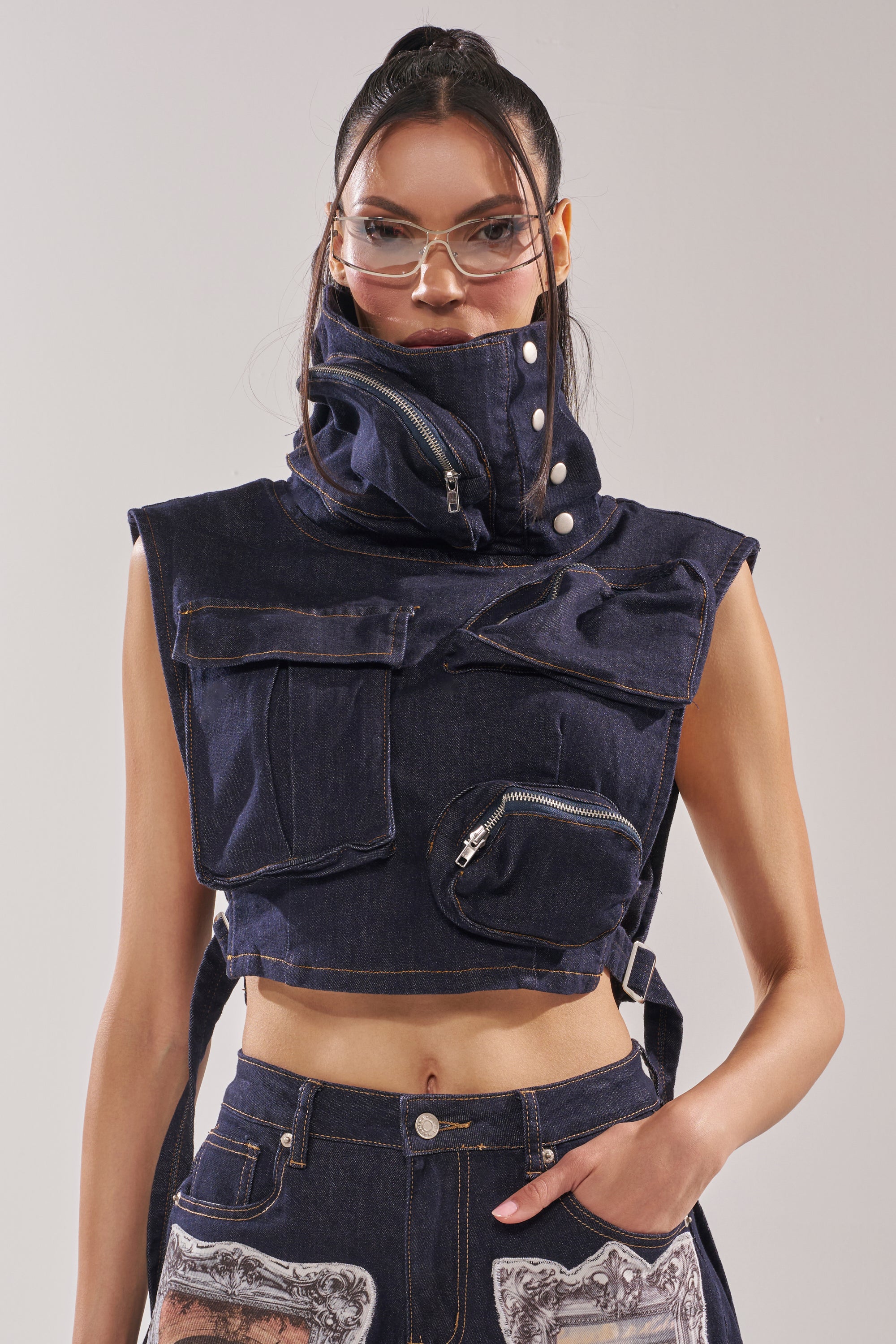 A woman wears the MODEL BEHAVIOR TOP IN DARK BLUE DENIM, featuring a large collar with zippered and snap details, styled with matching high-waisted jeans; she has long dark hair, clear glasses, and a neutral expression. 3