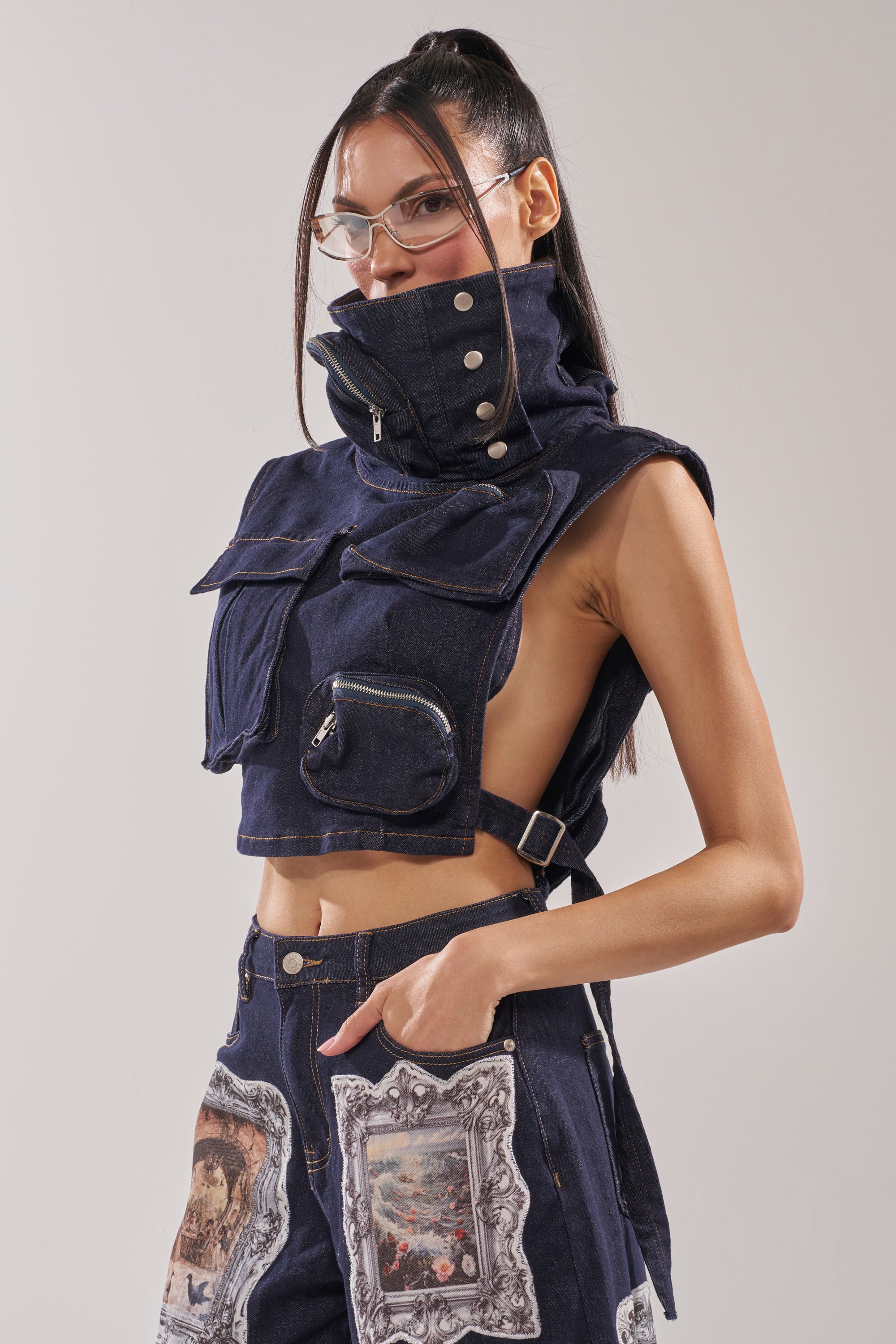 A woman wears the MODEL BEHAVIOR TOP IN DARK BLUE DENIM, a high-collared, sleeveless cargo-style vest with large pockets, metal buttons, and zippers. She pairs it with artistic print pants and clear glasses for a futuristic look. 4