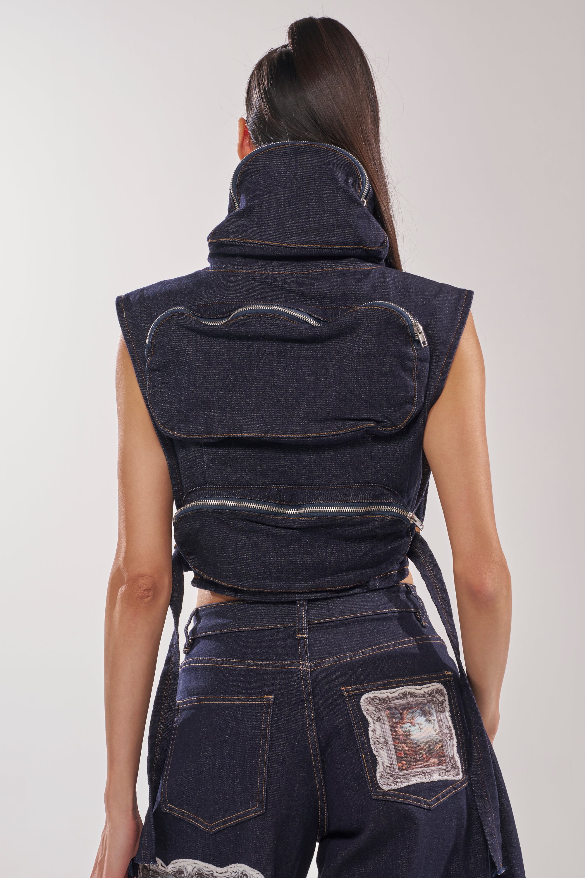 A person stands facing away, wearing the MODEL BEHAVIOR TOP IN DARK BLUE DENIM with large horizontal zippered pockets on the back and matching high-waisted jeans featuring patch artwork on the pocket. 6