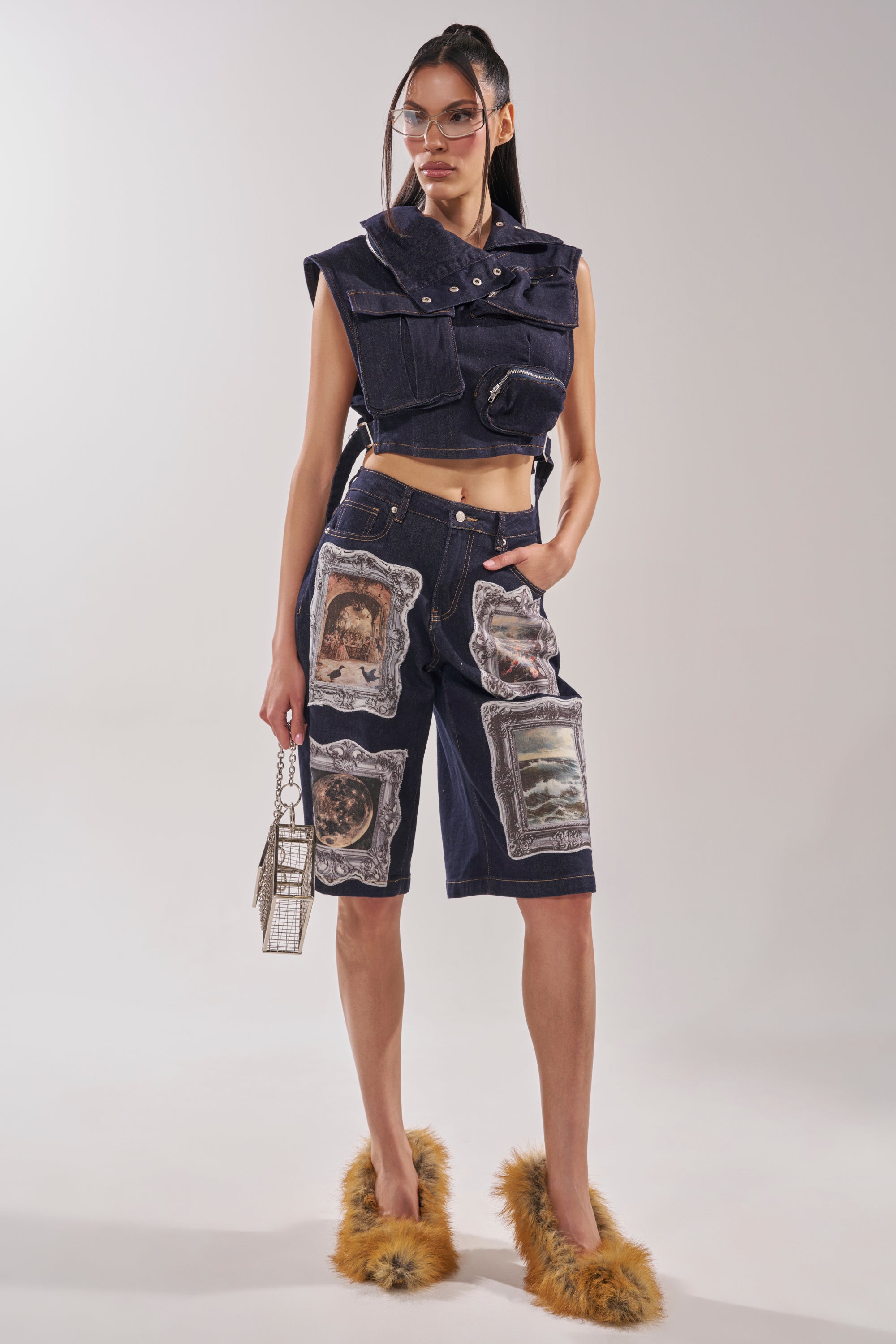 A woman wearing the MODEL BEHAVIOR TOP IN DARK BLUE DENIM, dark denim shorts with printed images, a small cage purse, clear glasses, and fluffy orange slippers stands against a plain light background. 7