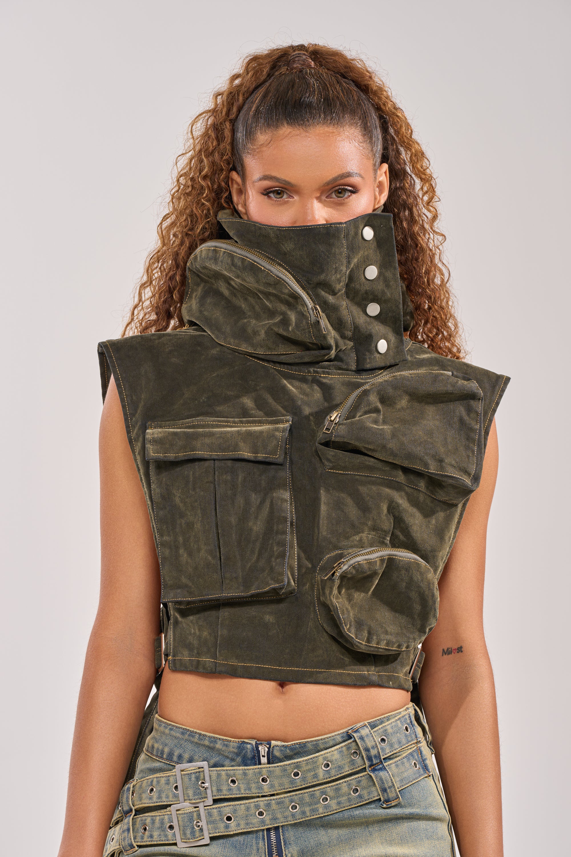 A woman with curly hair rocks a festival streetwear vibe, wearing the MODEL BEHAVIOR TOP IN DARK OLIVE DENIM and light blue jeans adorned with multiple belts and buckles. 0