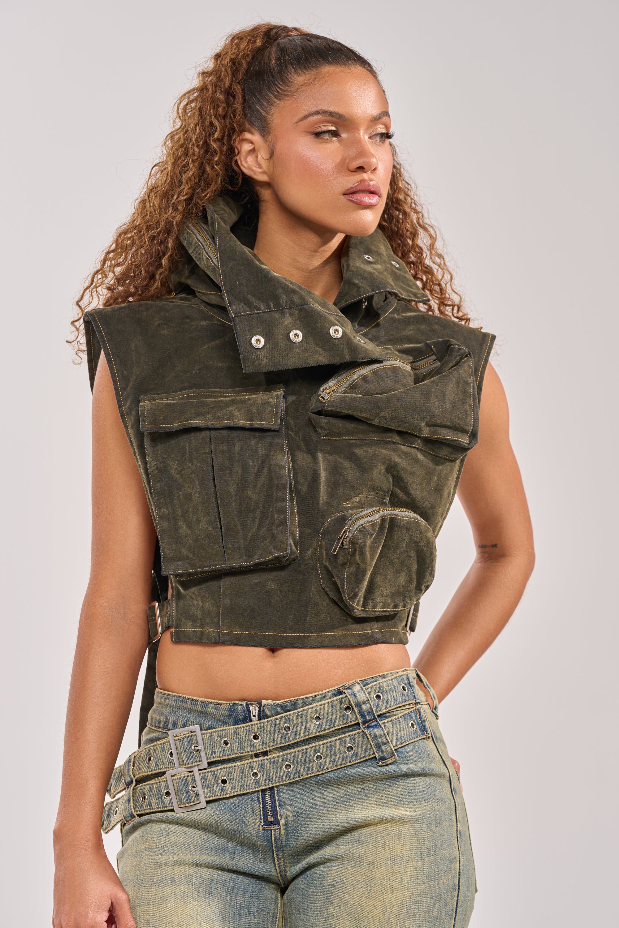 A woman with curly hair models the MODEL BEHAVIOR TOP IN DARK OLIVE DENIM, a utility vest with large pockets and high collar, styled with double-belted light blue jeans. She poses against a neutral background, showcasing festival-ready streetwear. 1
