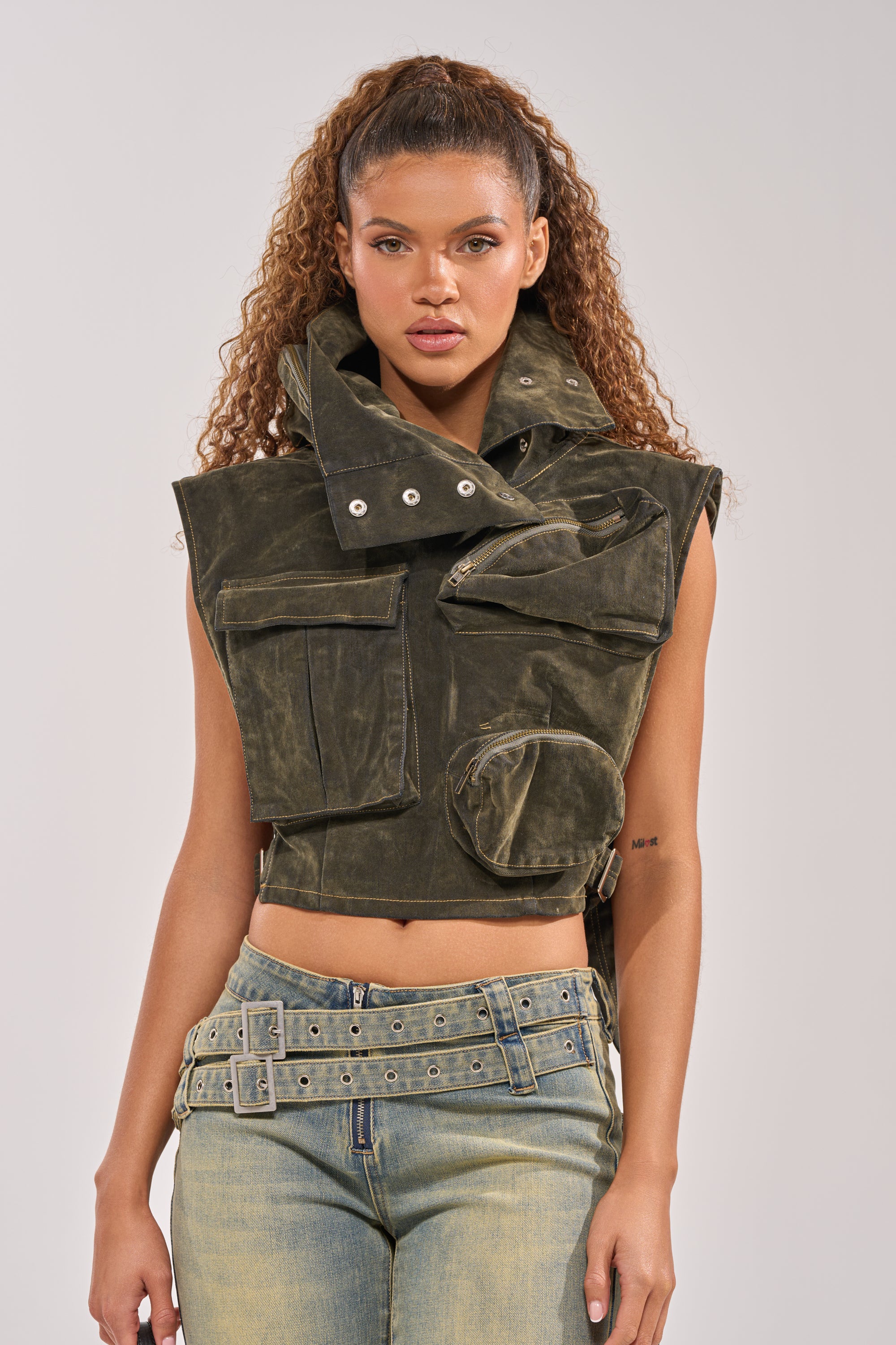 A woman stands facing forward against a plain background, wearing the MODEL BEHAVIOR TOP IN DARK OLIVE DENIM with multiple pockets and a large collar—effortlessly blending streetwear style with faded jeans and a double-buckle belt. 2