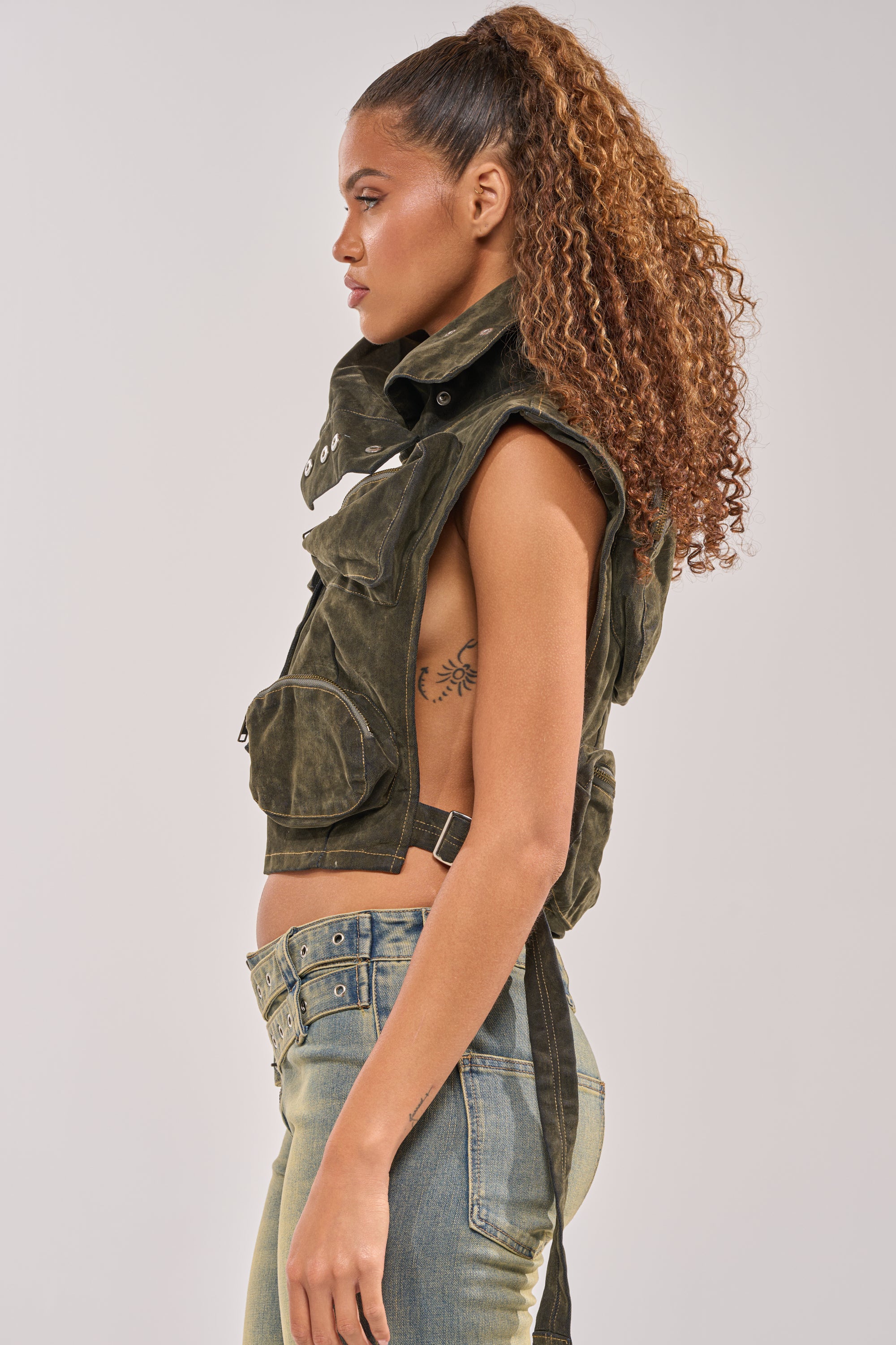 A young woman with curly hair in a high ponytail models the MODEL BEHAVIOR TOP IN DARK OLIVE DENIM and low-rise jeans, creating an effortless streetwear vibe as she stands in profile against a plain light gray background. 4