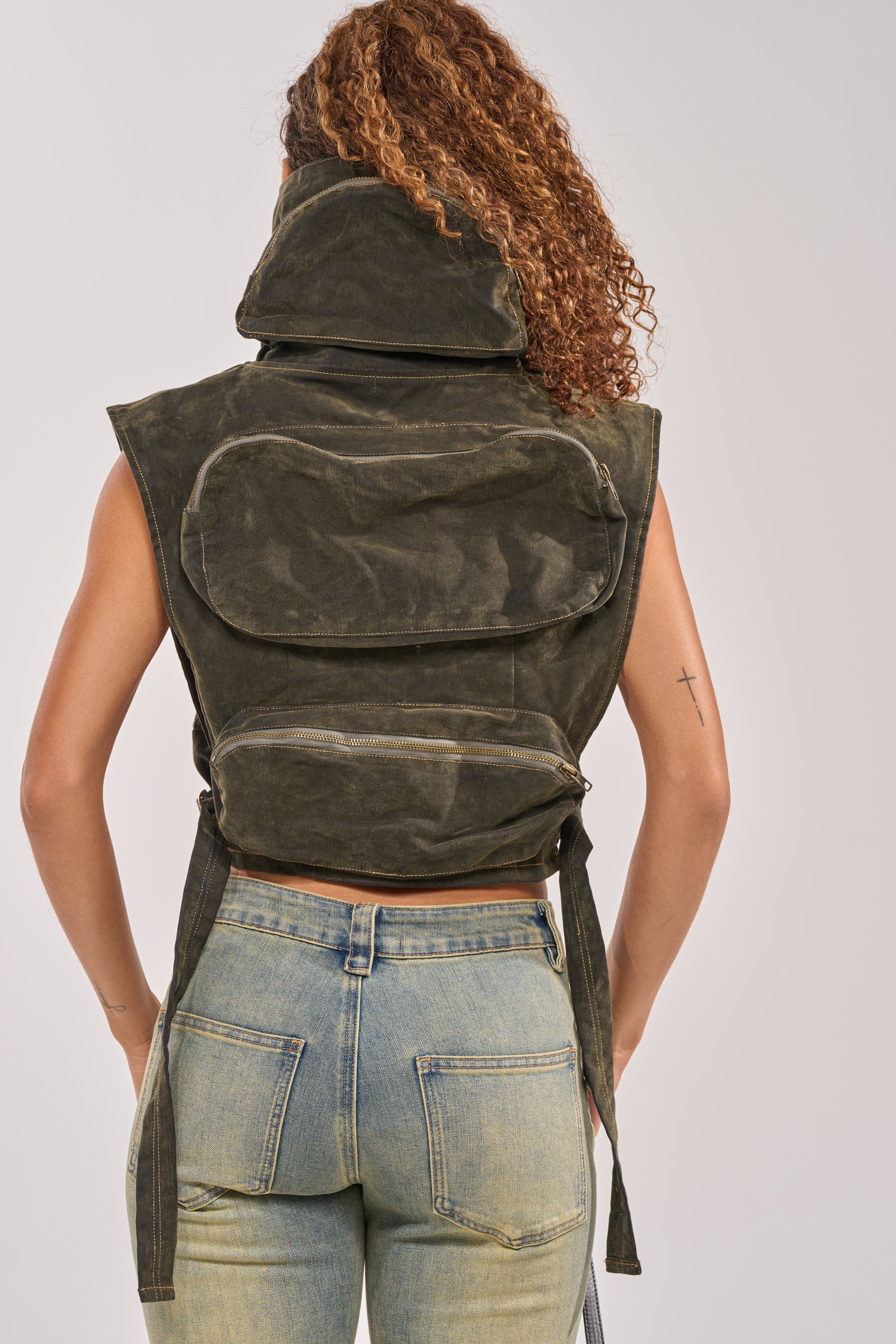 A person with curly hair wears the MODEL BEHAVIOR TOP IN DARK OLIVE DENIM— a sleeveless, high-collared cargo vest with zippered pockets—paired with faded light blue jeans for a streetwear vibe. A small cross tattoo is on their upper left arm. 5
