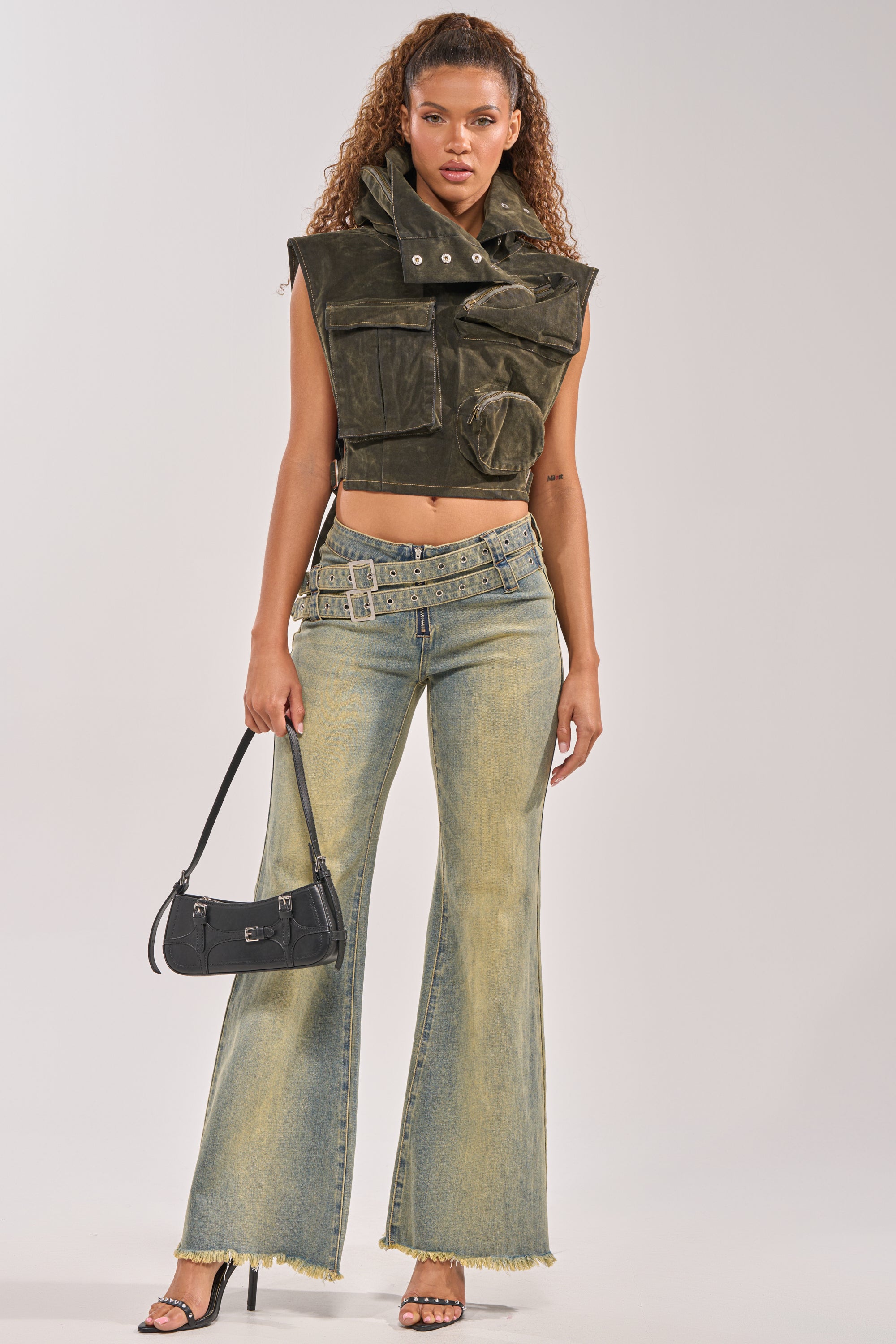 A woman faces forward wearing the Model Behavior Top in Dark Olive Denim, a sleeveless cargo vest with pockets and a high collar, styled with flared light-wash jeans, black heeled sandals, and a small black handbag. 6