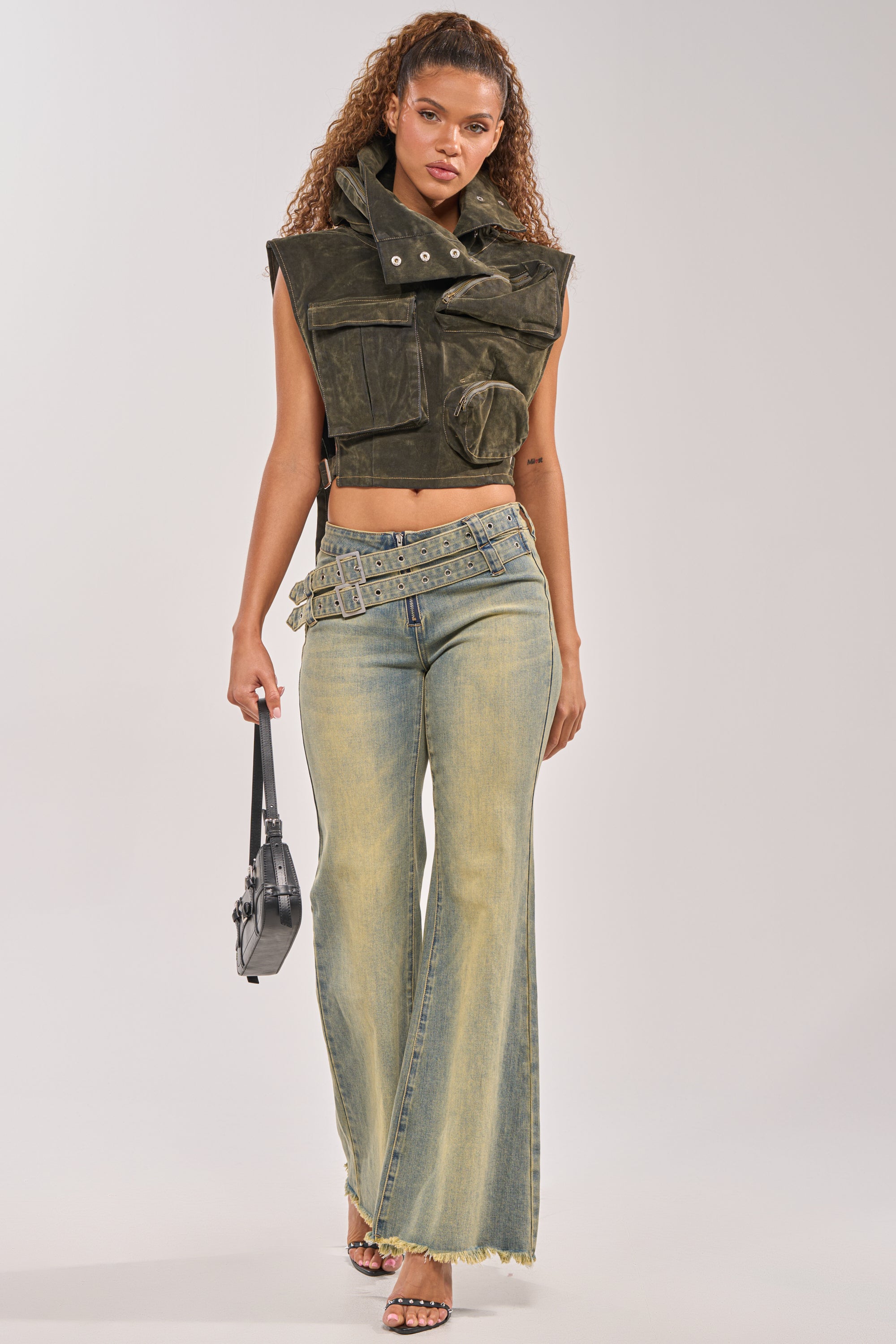 A woman with curly hair rocks festival streetwear in the MODEL BEHAVIOR TOP IN DARK OLIVE DENIM, paired with wide-leg, light-wash jeans, a loose belt, a small black handbag, and open-toe heels. 7