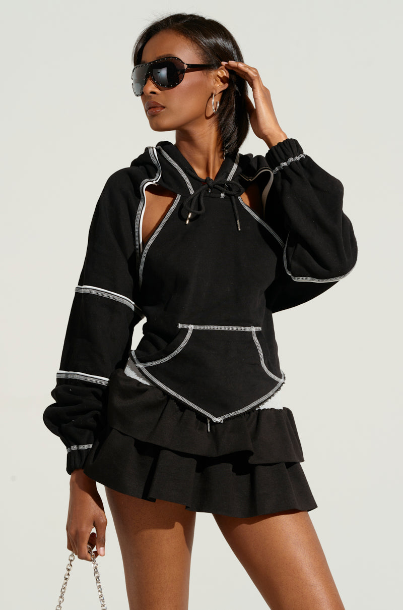 A person confidently dons a stylish black ensemble featuring the "ON A LIGHT DAY CUT OUT TIE BACK SWEATSHIRT" with its cropped silhouette and unique cut-out details. They pair large sunglasses and hoop earrings with the outfit, perfectly complemented by a chain-strap bag. The chic look is further accentuated by the tiered skirt.