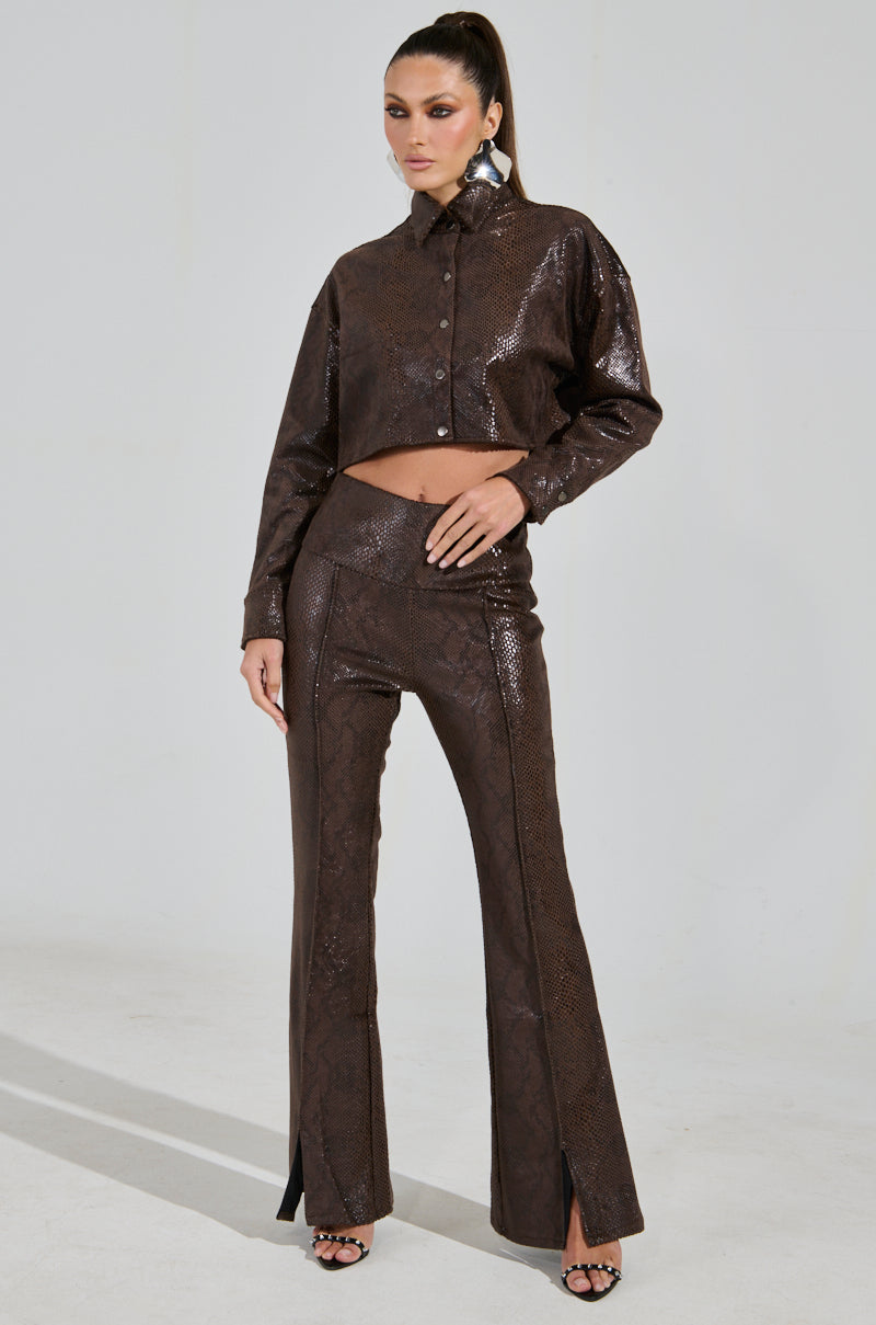 A model confidently poses in a shiny, dark brown two-piece outfit with a cropped button-up top and DROP IT LOW PANT WITH SLIT. She stands with one hand on her hip, wearing high heels in a well-lit studio.