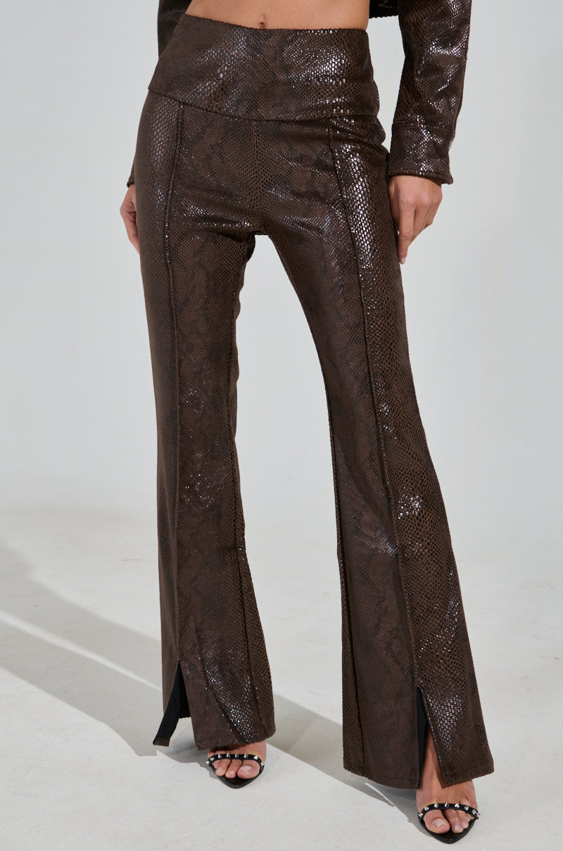 A person flaunts the DROP IT LOW PANT WITH SLIT, featuring a high-rise snake-print and shiny faux leather texture with small front slits at the hems. Paired elegantly with stylish black open-toe heels, the photo captures chic lower body fashion.