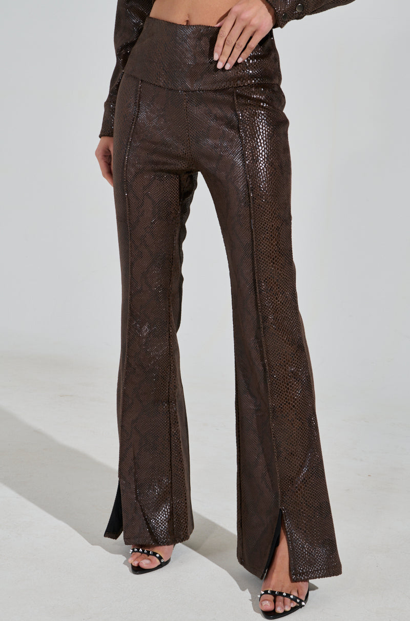 A person wears the DROP IT LOW PANT WITH SLIT, featuring brown high-rise faux leather with a sleek snakeskin pattern and front slits. They pair it with black open-toed high heels against a plain, light studio backdrop.