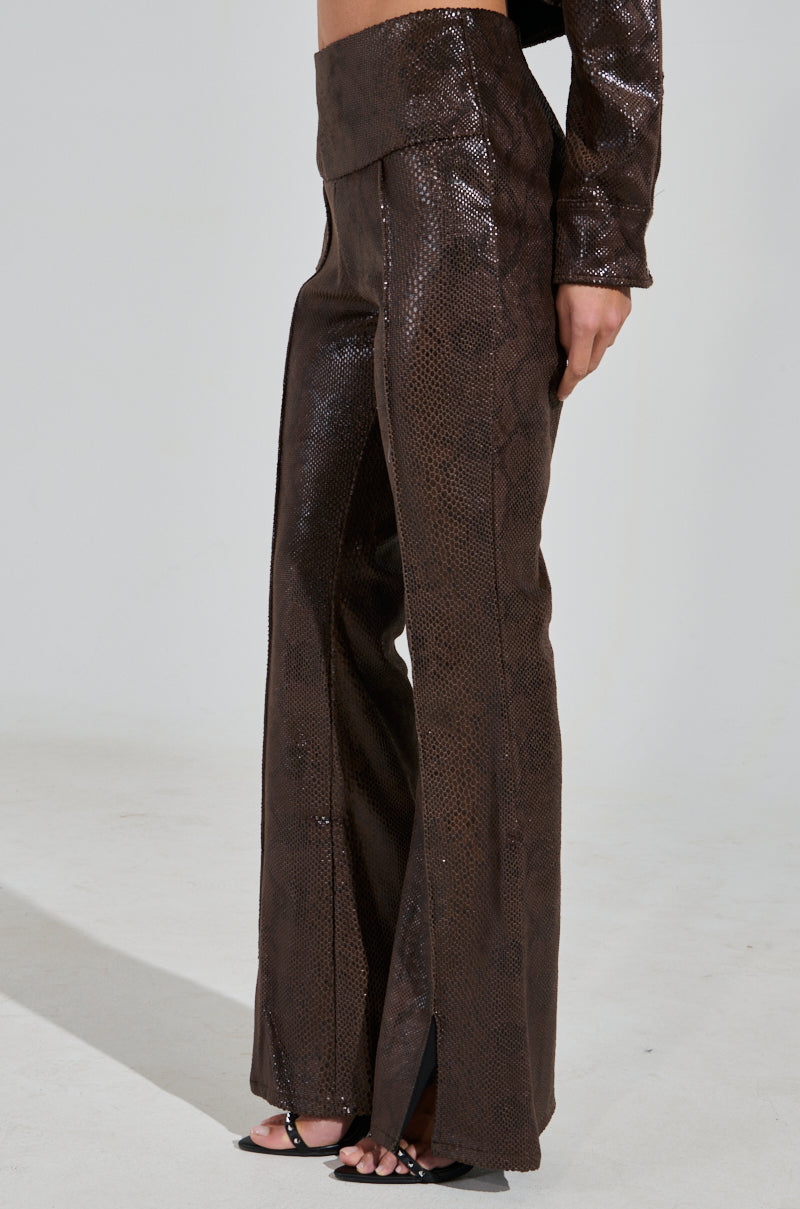 Wearing the DROP IT LOW PANT WITH SLIT, featuring a high-rise snake pattern and shimmering faux leather, paired with matching top fabric. A hand rests by their side against a plain light background, exuding chic elegance.