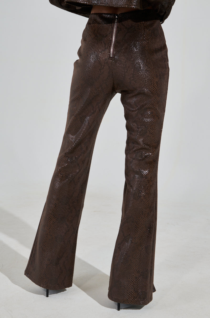Wearing brown, crocodile-patterned DROP IT LOW PANT WITH SLIT trousers, a person stands facing away from the camera. The faux leather pants have a glossy finish with a visible back zipper, set against a plain background.