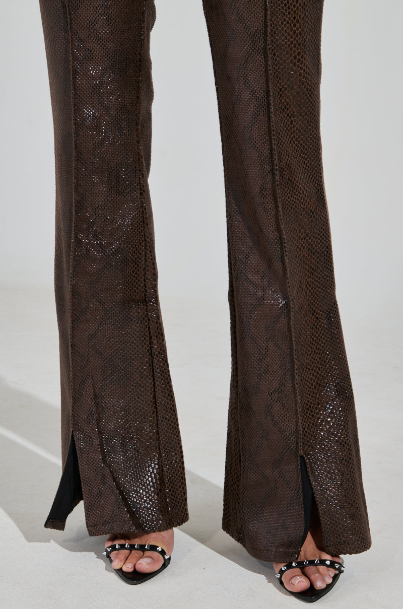 Wearing the DROP IT LOW PANT WITH SLIT, these brown faux leather pants feature a snakeskin pattern with ankle slits. Paired with black strappy high-heeled sandals adorned with silver studs, standing on a light floor.