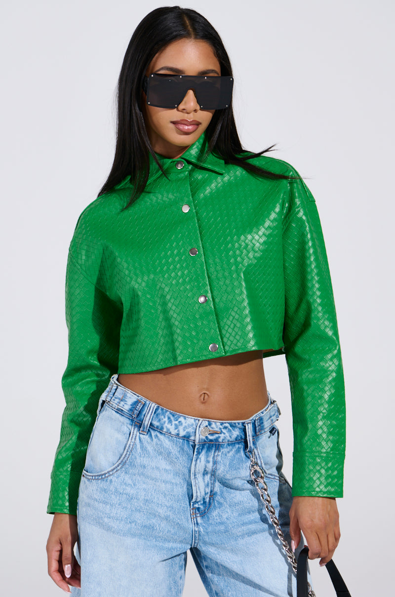 A woman wears oversized sunglasses and a shiny green TOO EXPENSIVE FOR YOU WOVEN PU CROPPED BUTTON UP SHIRT with silver buttons. She pairs it with light blue denim jeans featuring a chain detail, exuding confidence in her bold style.