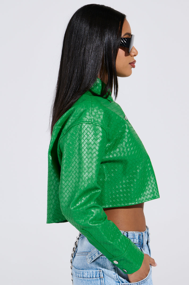 Someone with long dark hair dons a bright green textured jacket over light blue jeans, with sunglasses on. They're turned sideways against a plain backdrop, revealing the TOO EXPENSIVE FOR YOU WOVEN PU CROPPED BUTTON UP SHIRT subtly beneath their vibrant outerwear.