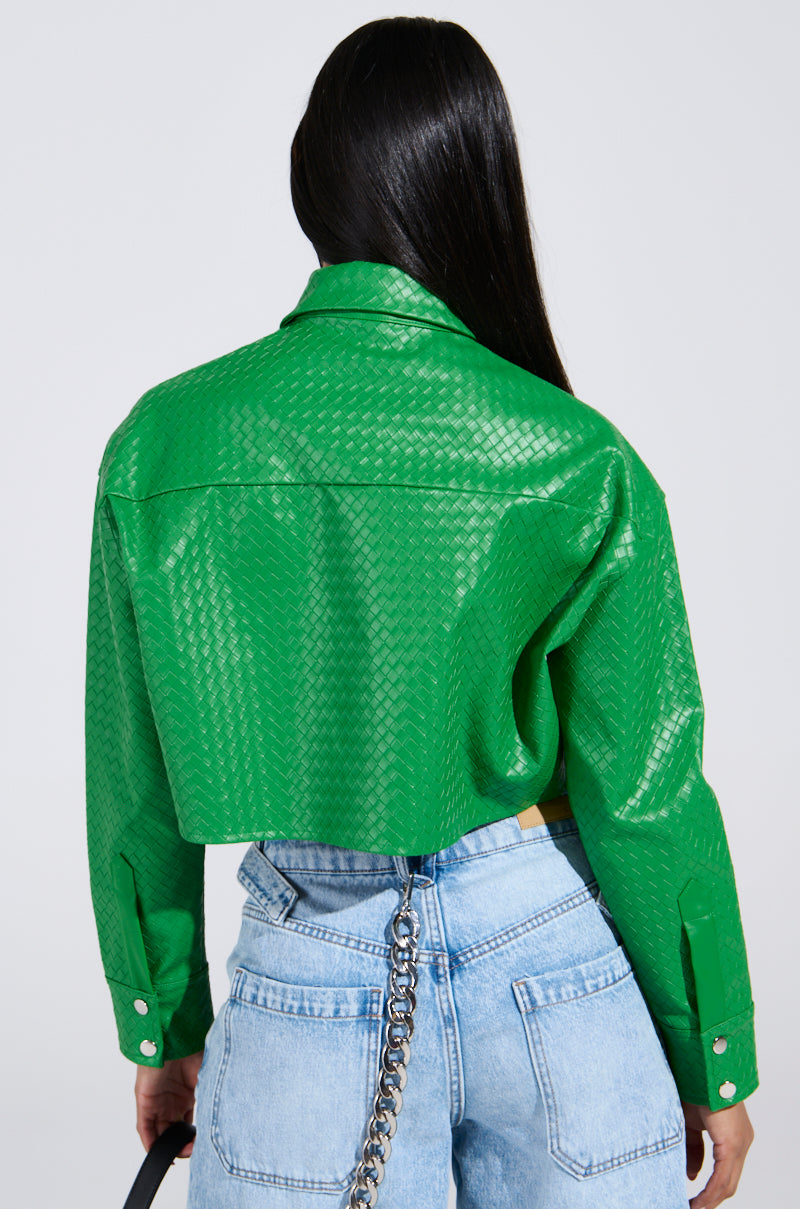 With long dark hair, a person wears the "TOO EXPENSIVE FOR YOU" bright green textured cropped button-up jacket and light blue jeans with a chain accessory. Seen from the back against a plain background, they hold a small black handbag in their right hand.