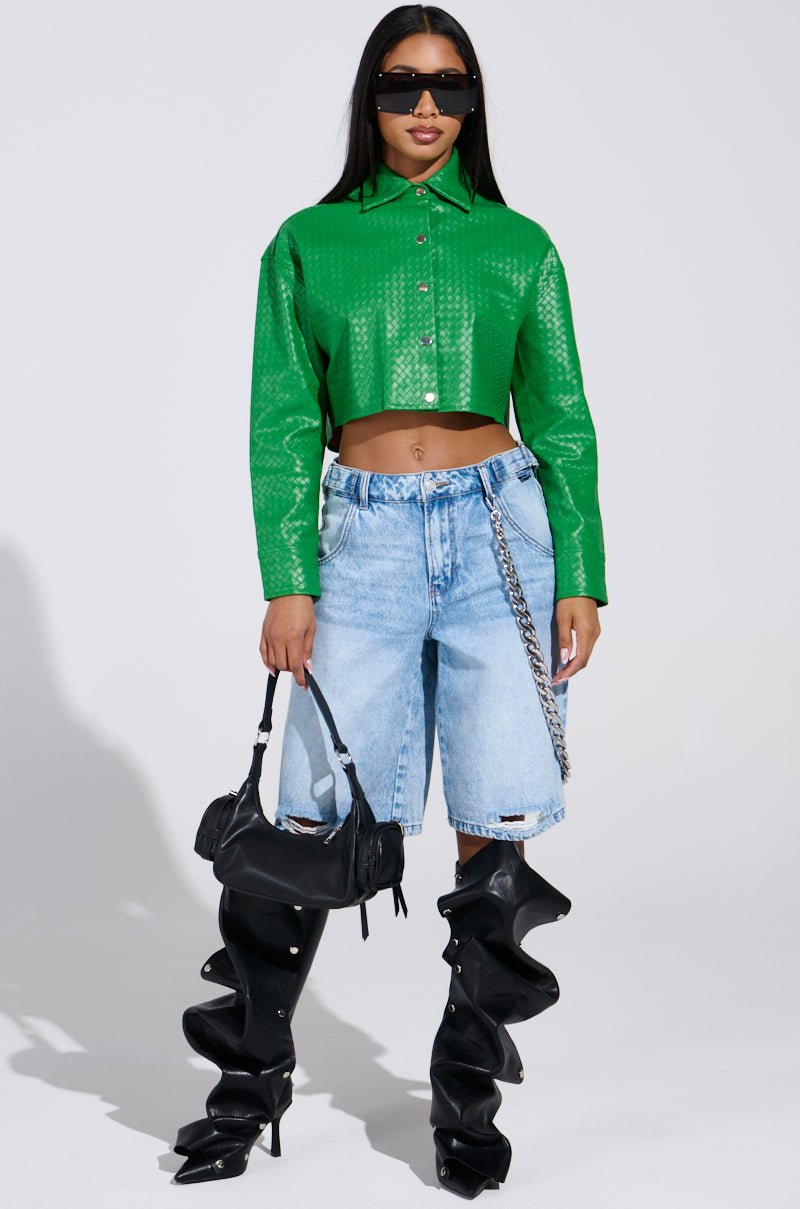 A person in a green, long-sleeve crop top and light denim shorts holds a black handbag. They're wearing black, futuristic high-heeled boots and large sunglasses, standing against a plain backdrop with the chic vibe of the "TOO EXPENSIVE FOR YOU WOVEN PU CROPPED BUTTON UP SHIRT.