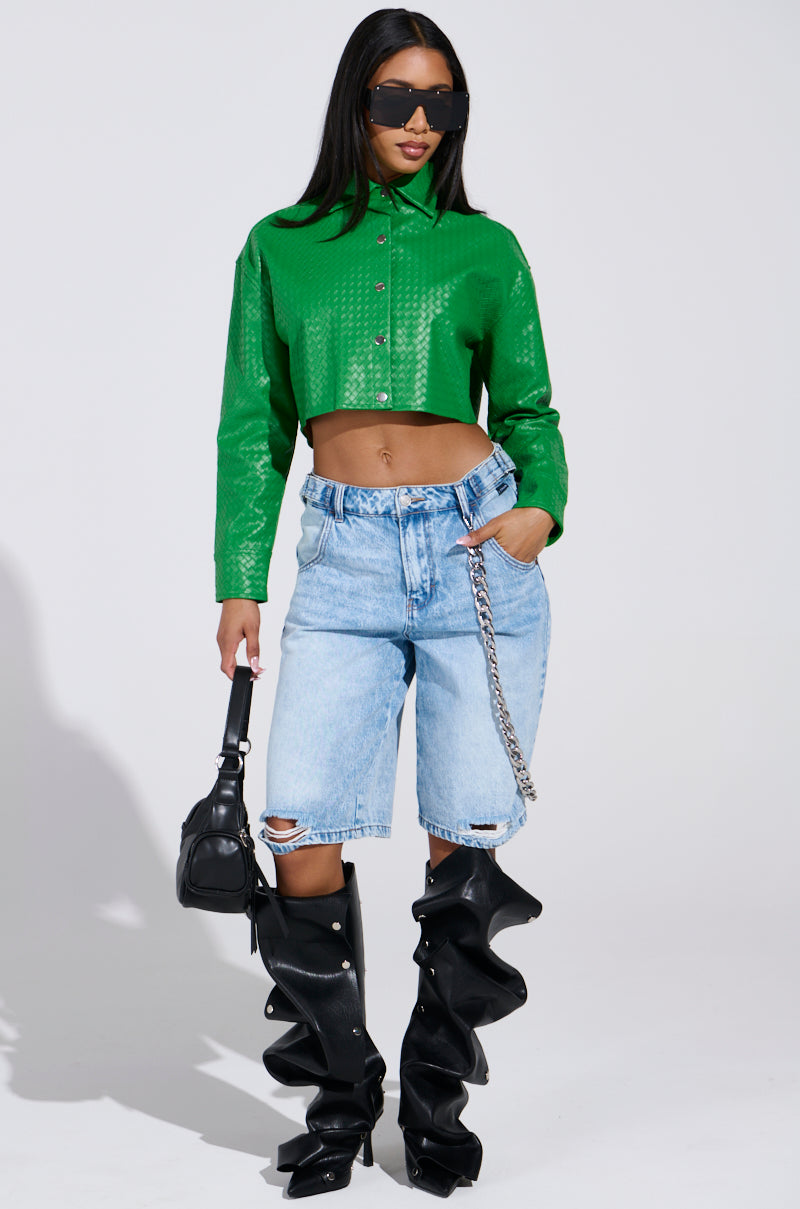 A woman in a green snakeskin-patterned TOO EXPENSIVE FOR YOU WOVEN PU CROPPED BUTTON UP SHIRT and light blue ripped denim shorts poses with one hand in her pocket, wearing oversized black sunglasses, black wavy thigh-high boots, and carrying a small black handbag.
