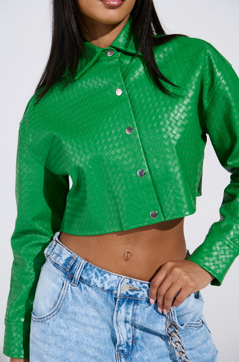 A person wears the TOO EXPENSIVE FOR YOU WOVEN PU CROPPED BUTTON UP SHIRT in vibrant green, paired with light blue jeans and a chain accessory, emphasizing the jacket's textured design and casually stylish look.