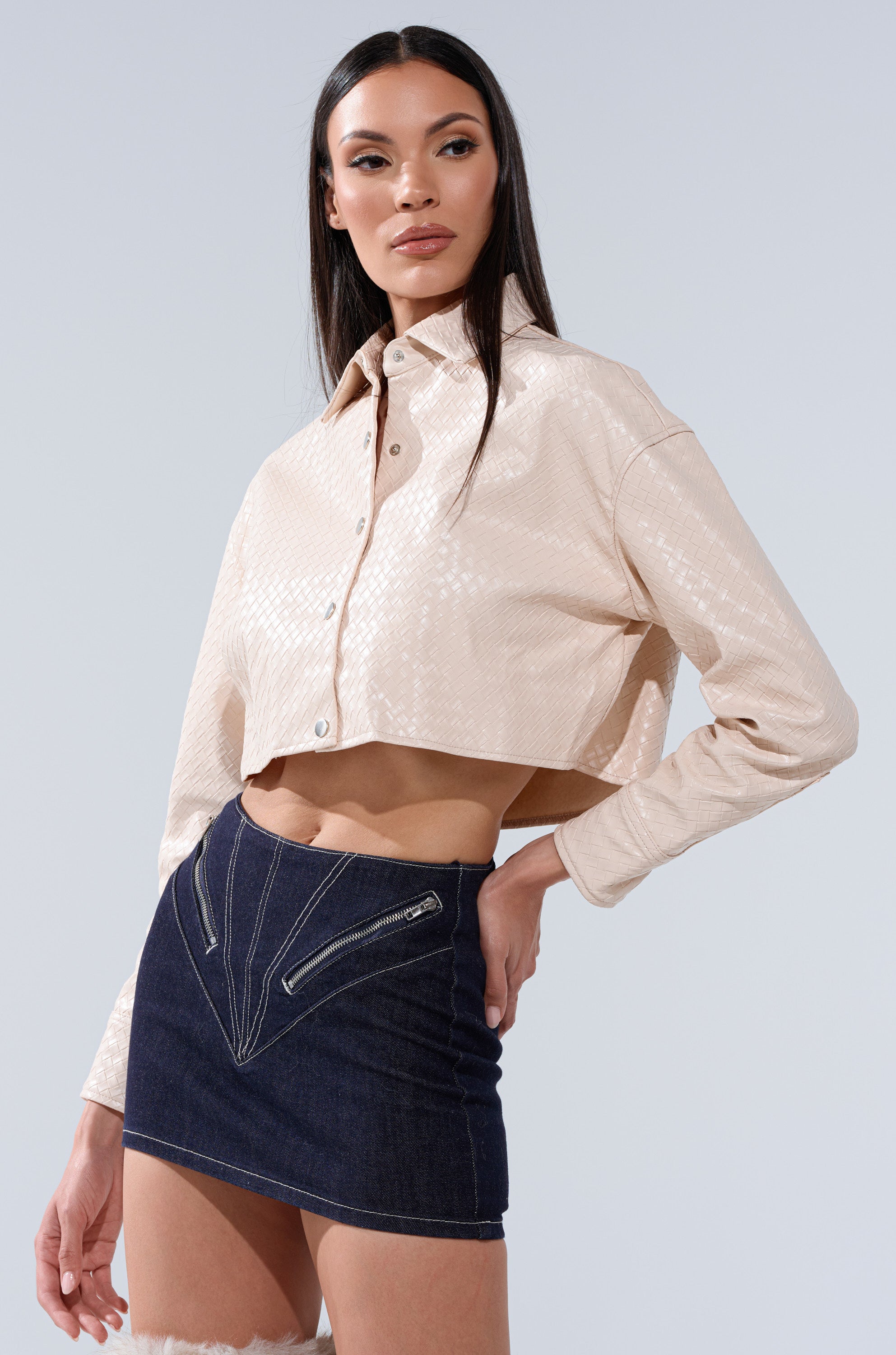 A woman poses against a plain light background wearing the LUXURY GIRL WOVEN PU CROPPED BUTTON UP SHIRT IN IVORY, paired with a high-waisted dark denim mini skirt featuring front zipper details.