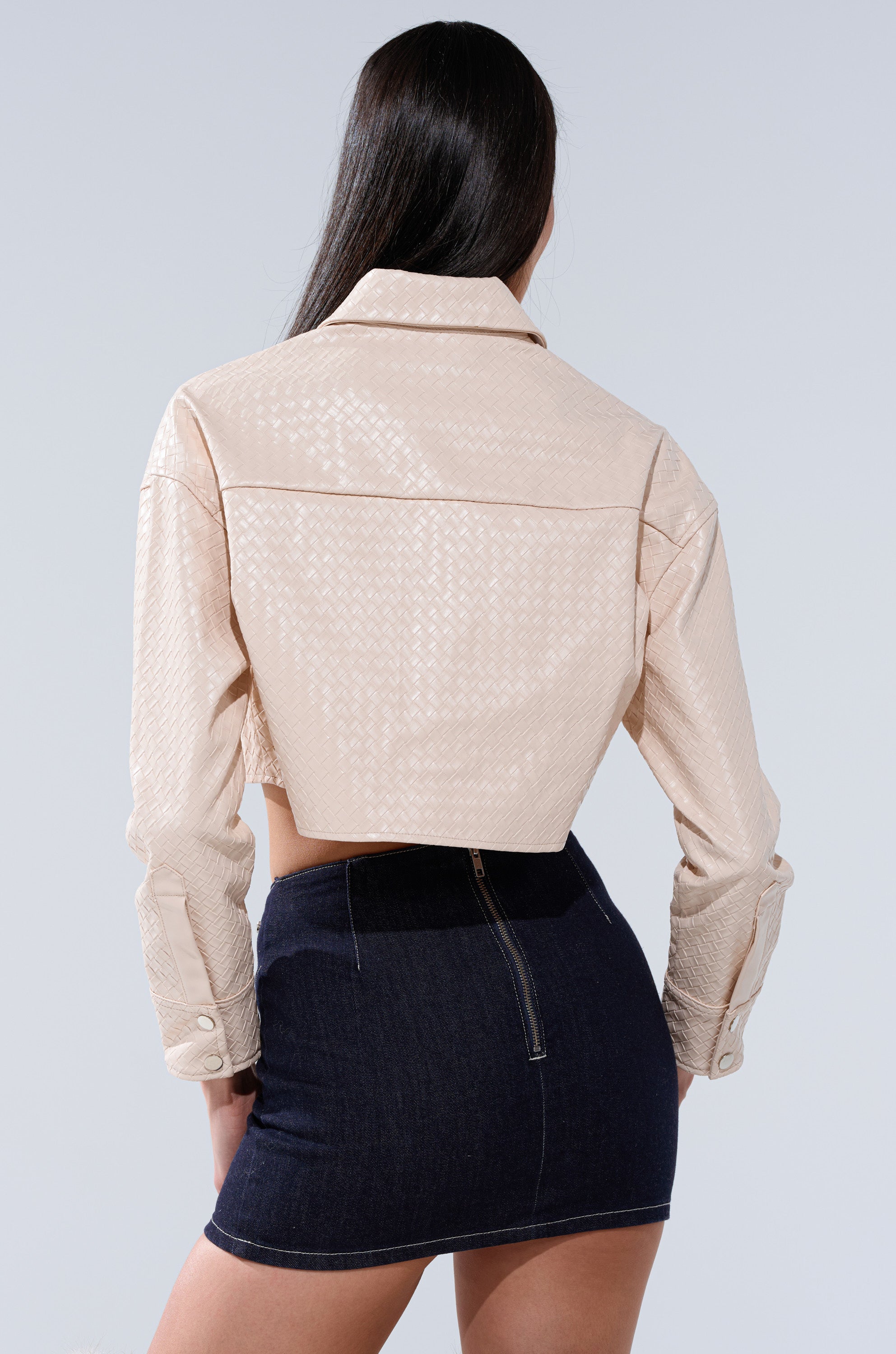 A woman stands facing away, wearing the LUXURY GIRL WOVEN PU CROPPED BUTTON UP SHIRT IN IVORY paired with a dark denim mini skirt featuring a visible back zipper, set against a plain light background.