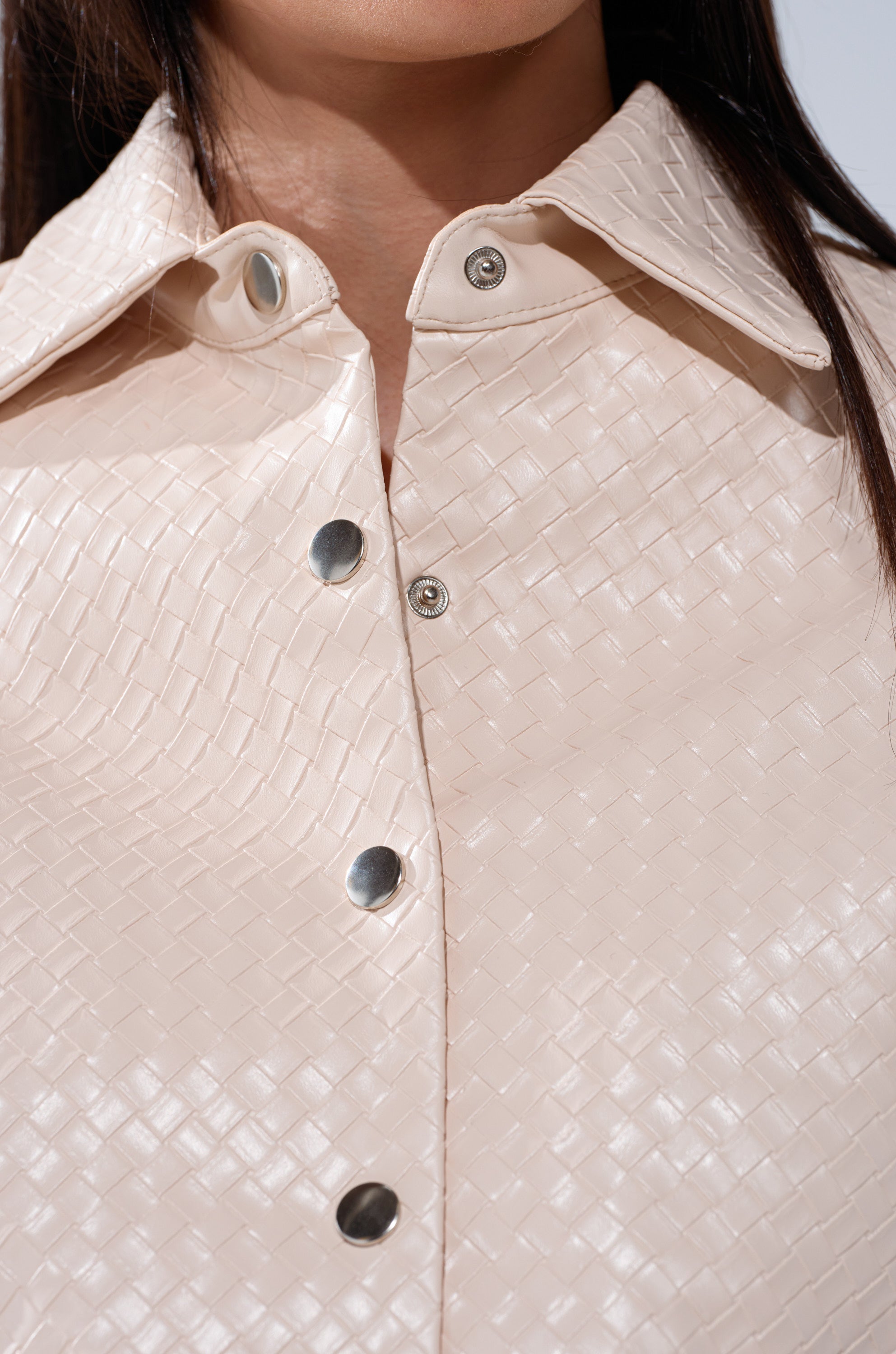 Close-up of a person wearing the LUXURY GIRL WOVEN PU CROPPED BUTTON UP SHIRT IN IVORY, featuring a boxy fit, woven pattern, metallic snap buttons, collar, and a textured finish. The person's dark straight hair is partially visible.