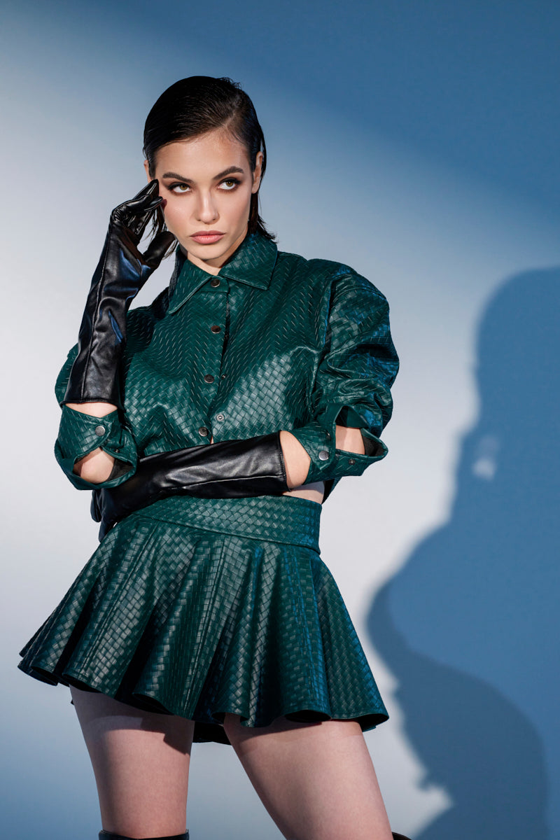 A woman poses in the LUXURY GIRL WOVEN PU CROPPED SHIRT IN GREEN with matching pleated skirt and long black gloves, standing against a blue background under dramatic lighting, looking to the side with one hand touching her head.