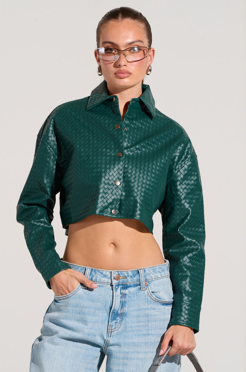 A person sporting the LUXURY GIRL WOVEN PU CROPPED SHIRT IN GREEN and light blue jeans stands against a plain background. Their hair is tied back, they are wearing glasses, and have one hand in their pocket.