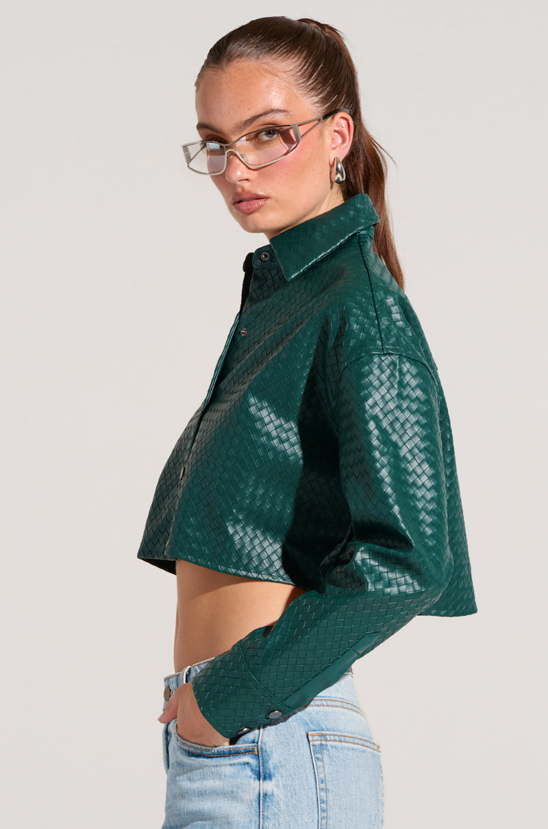 A person wearing a LUXURY GIRL WOVEN PU CROPPED SHIRT IN GREEN and light blue jeans stands in profile. They have long hair tied back and wear rectangular glasses. The background is plain and light-colored.