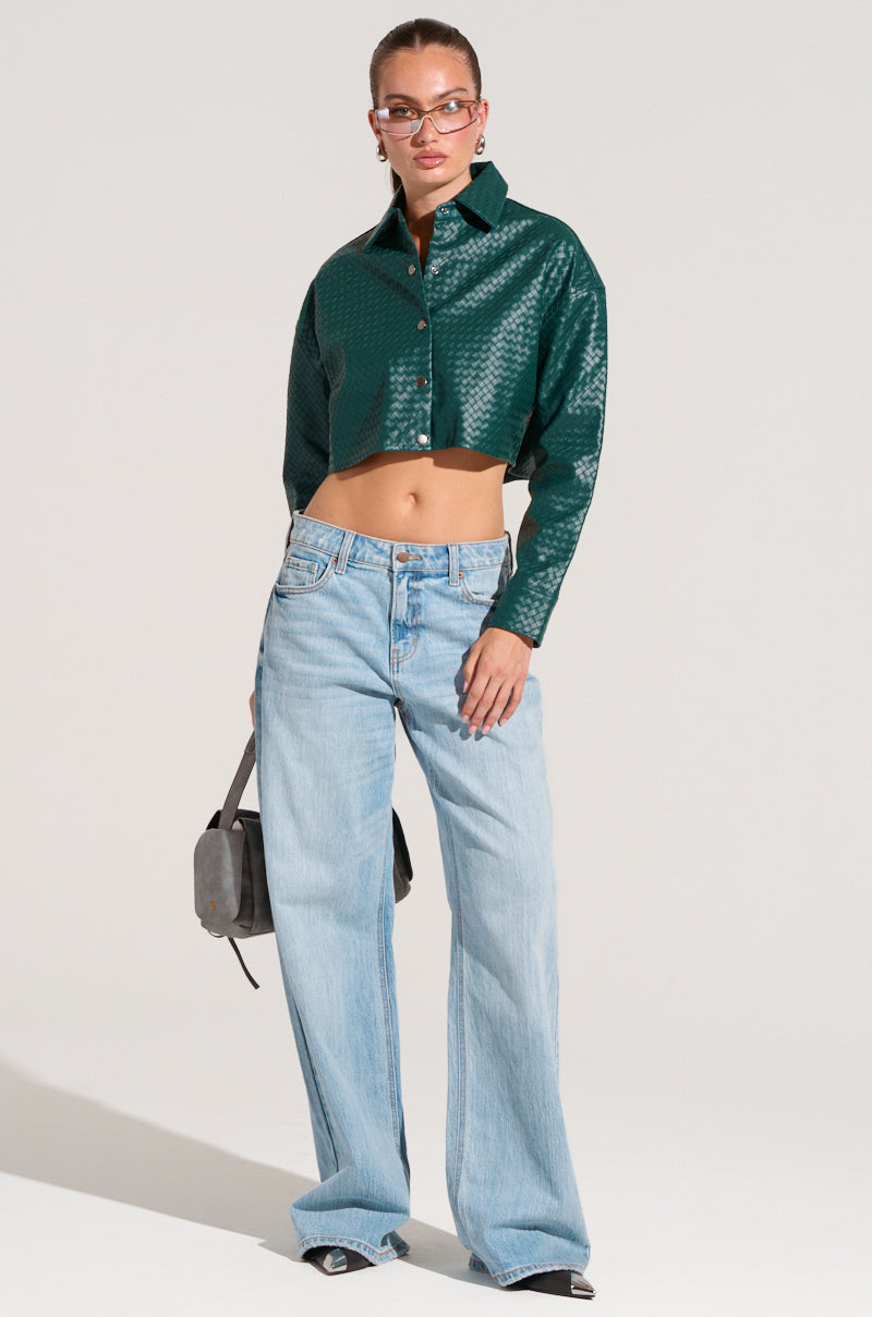 A person stands against a plain background, wearing the LUXURY GIRL WOVEN PU Cropped Shirt in Green, light blue high-waisted wide-leg jeans, and black shoes. They are accessorized with sunglasses and hold a gray handbag in their left hand.