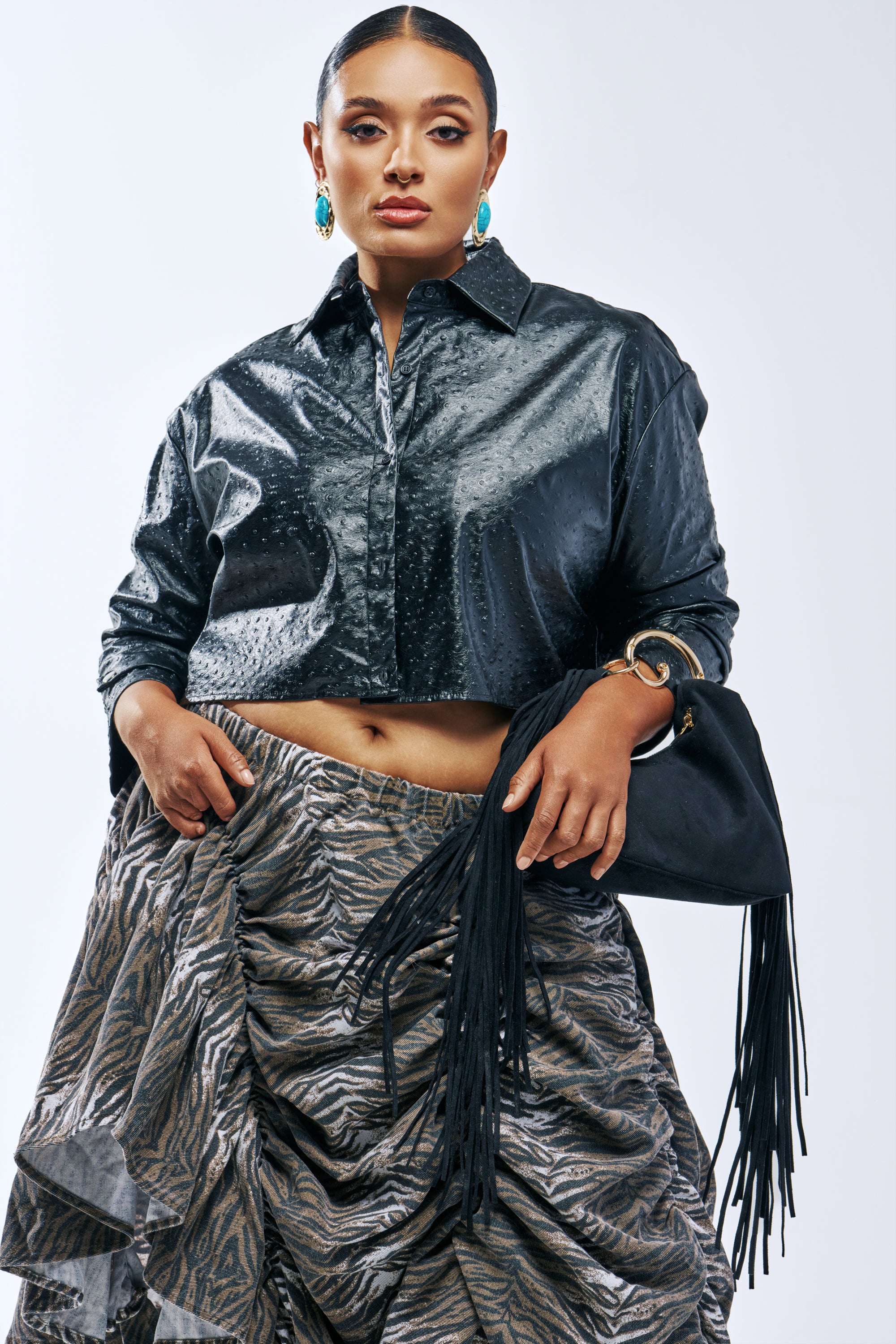 A person wears a black TOO GOOD FOR YOU FAUX OSTRICH LEATHER BUTTON DOWN, a ruched skirt with a grey and brown animal print, and carries a black handbag with long fringe.