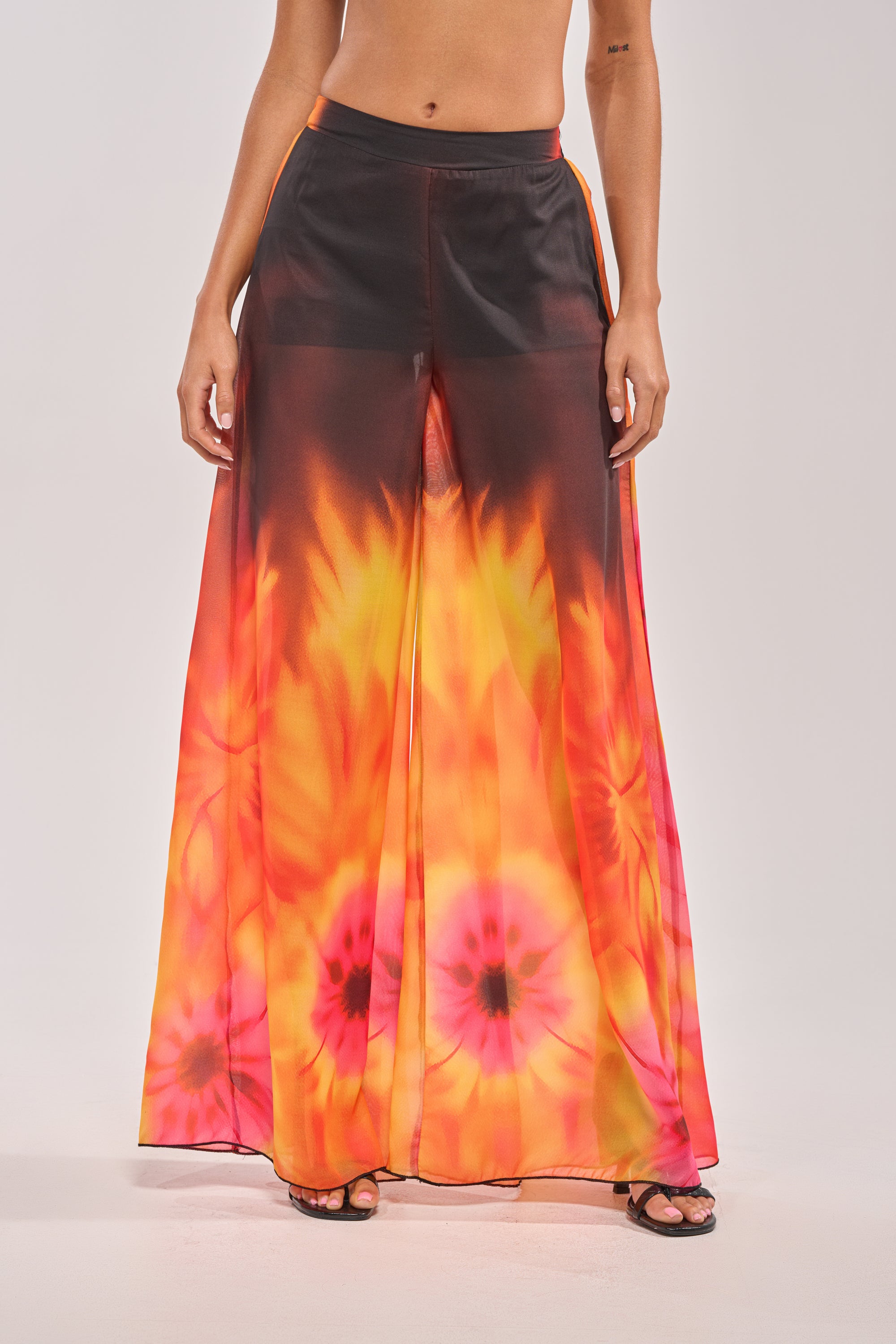 A person styles the GIRL ON FIRE CHIFFON PALAZZO PANT—sheer, high-waisted, wide-leg floral pants in vibrant orange, yellow, and pink—with black sandals for a chic look.