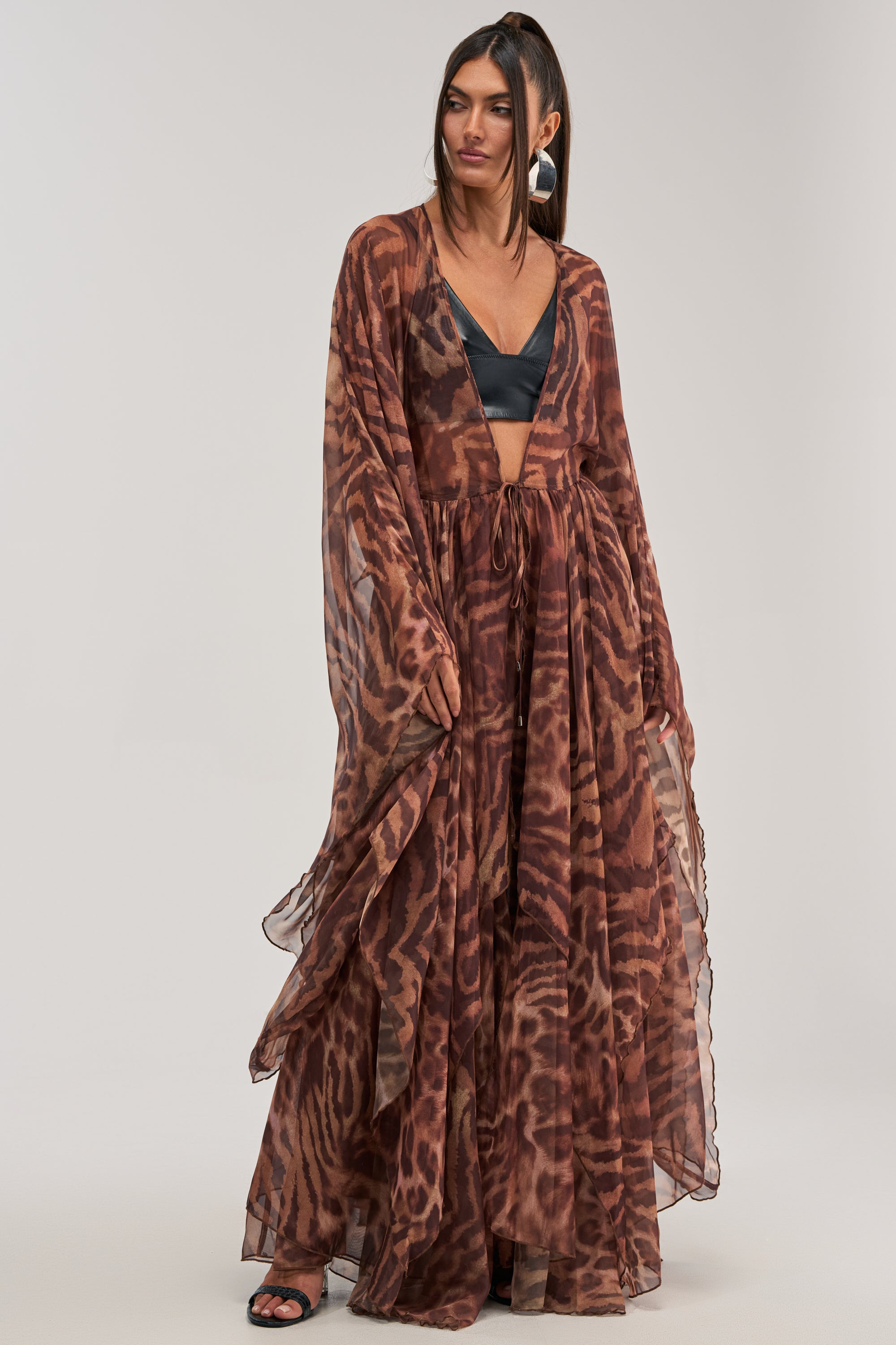 A woman stands against a plain light background wearing the MOONLIGHT TIGER DUSTER, a flowing brown tiger print chiffon maxi dress with a deep V-neck and sheer long sleeves, styled with a black bralette and open-toe black sandals.