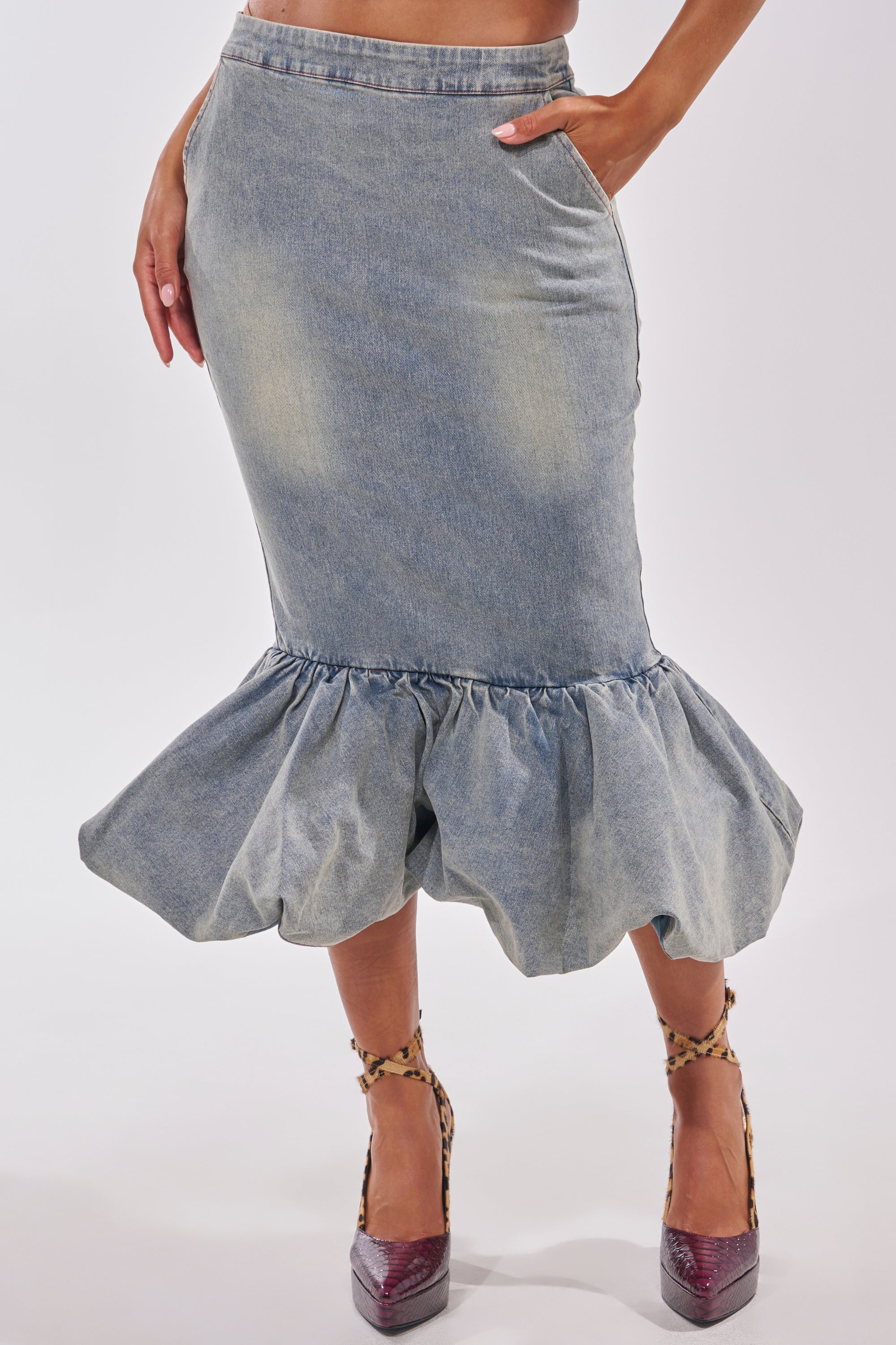 Wearing the JOSIE DENIM MIDI SKIRT, a figure poses with one hand on their hip, showcasing the curve-hugging fit and ruffled hem, paired with leopard-print ankle-strap heels against a plain white background. 1