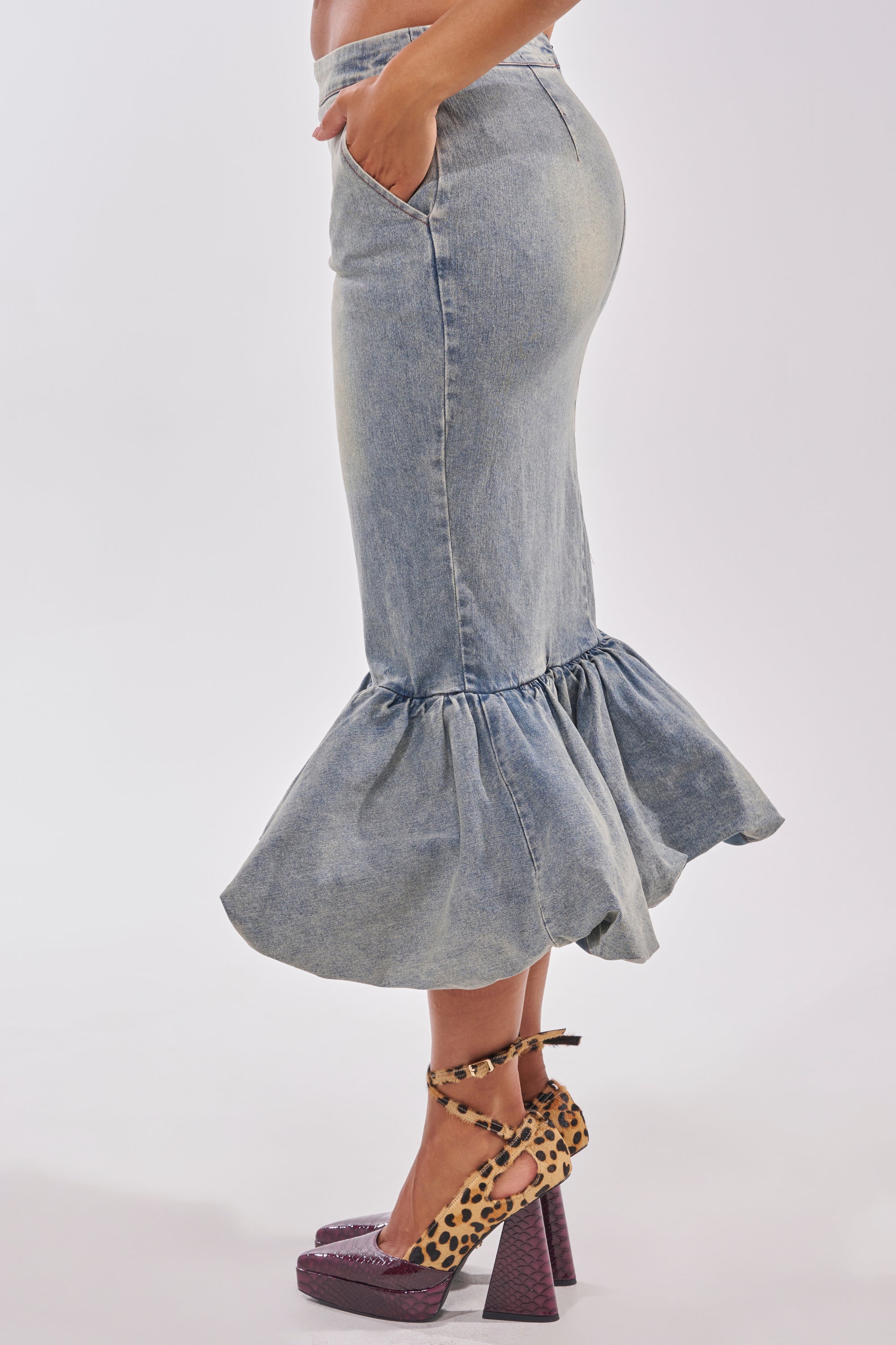 A person wears the curve-hugging JOSIE DENIM MIDI SKIRT with a flared hem in light wash, paired with leopard print ankle-strap platform heels, standing against a plain light background. 2