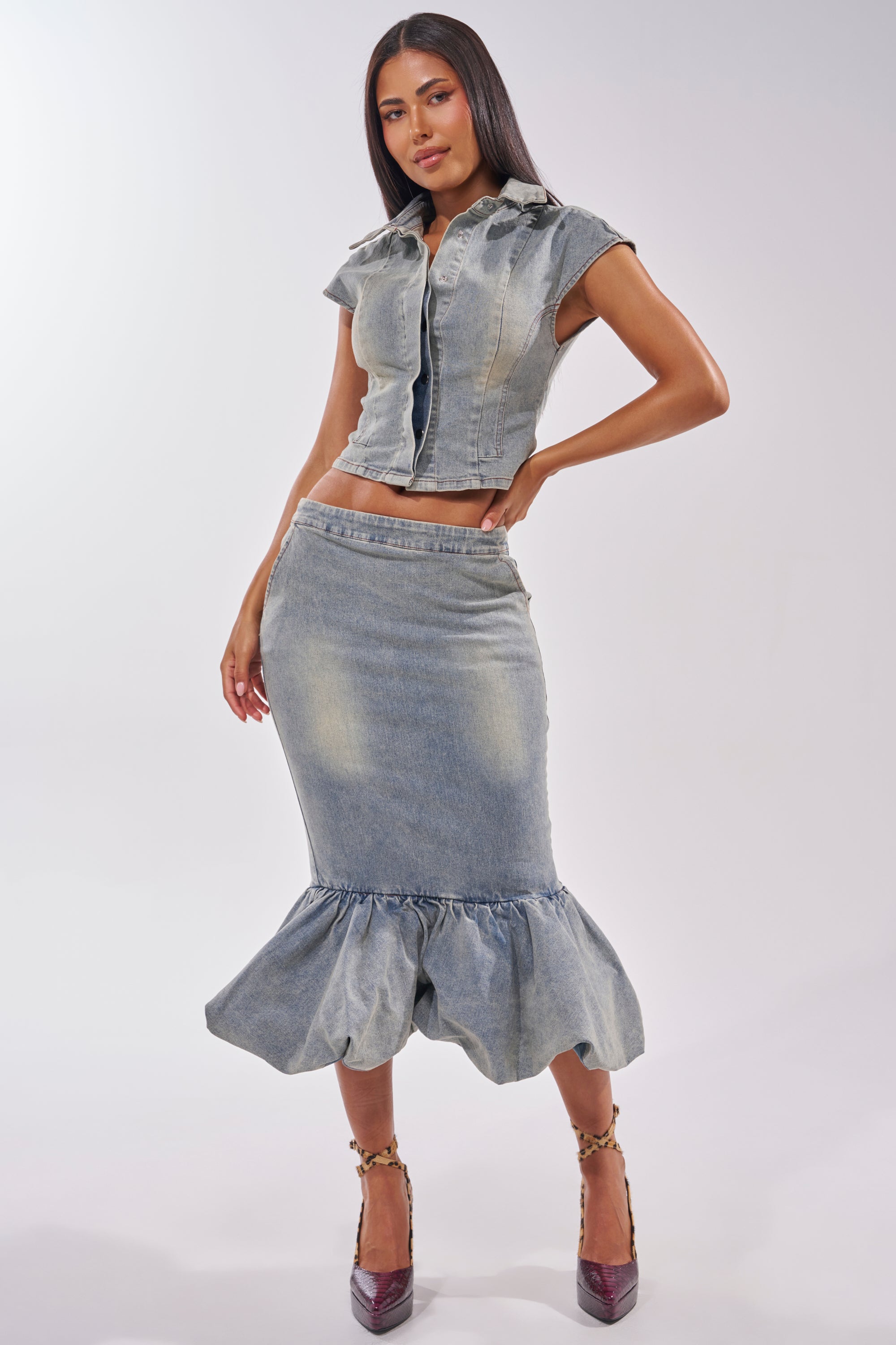 A woman models the JOSIE DENIM MIDI SKIRT, a curve-hugging denim piece with ruffled details, paired with a sleeveless cropped jacket and high heels against a plain light background. 6
