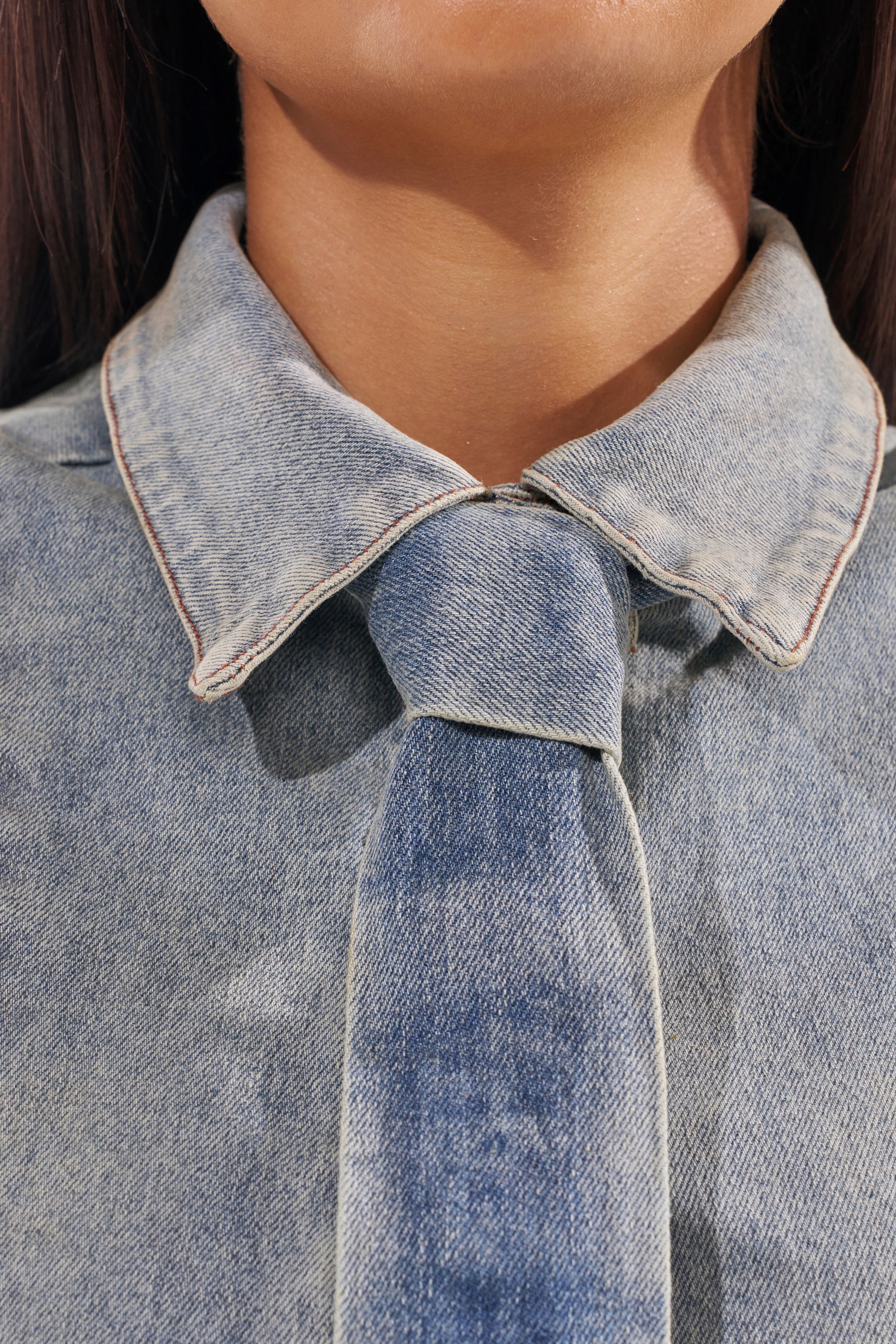 Close-up of a person wearing the JOSIE DENIM BLOUSE with a matching denim tie, both featuring visible stitching. Focus is on the collar and upper chest, highlighting this classic statement piece; the face is partially out of frame. 10