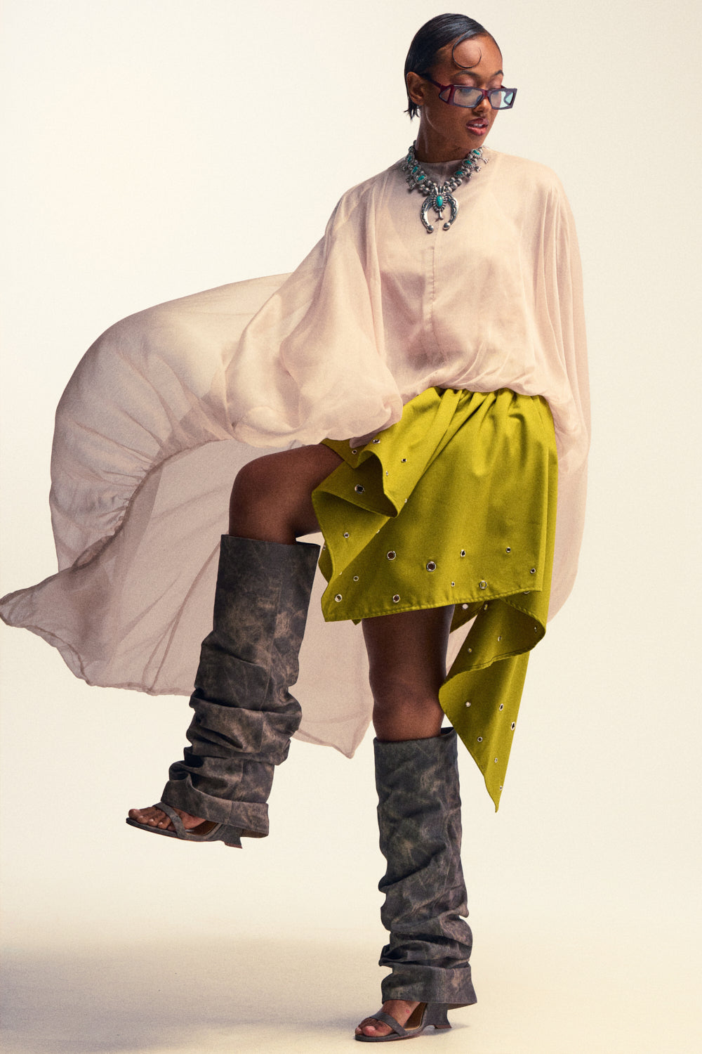 A person poses confidently against a plain light background, wearing the HEAD IN THE CLOUDS CHIFFON BLOUSE—an ivory blouse with a flowing chiffon fabric and draped silhouette—paired with a bright yellow embellished skirt, layered necklaces, statement glasses, and tall slouchy gray boots.