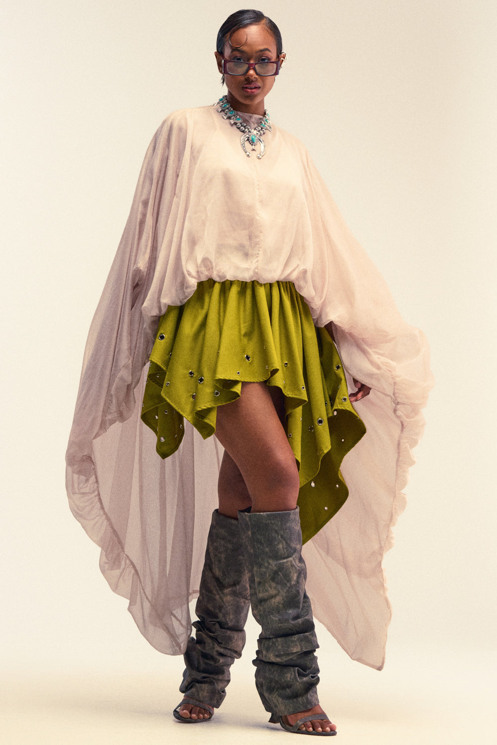 Wearing the "HEAD IN THE CLOUDS CHIFFON BLOUSE," a flowing pale pink high-low top, paired with a bright green embellished skirt, tall gray slouch boots, a statement necklace, and sunglasses, a person poses confidently against a light background.