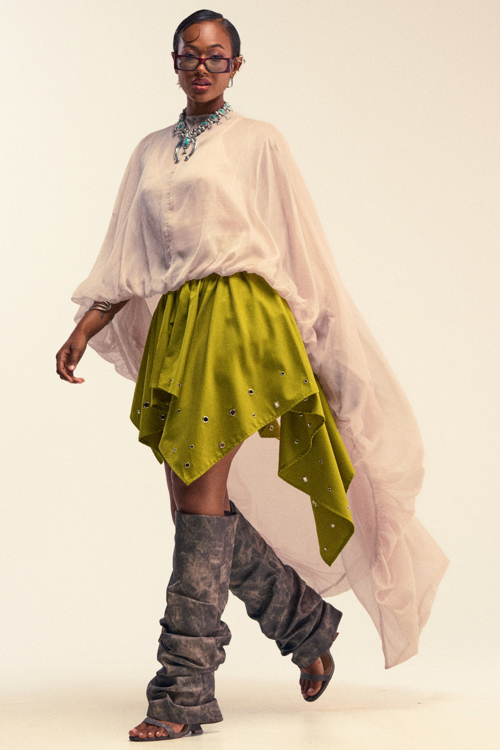 Wearing the HEAD IN THE CLOUDS CHIFFON BLOUSE, a woman pairs it with an olive green asymmetrical embellished skirt, tall grey slouchy boots, a bold necklace, and glasses as she walks confidently against a light background.